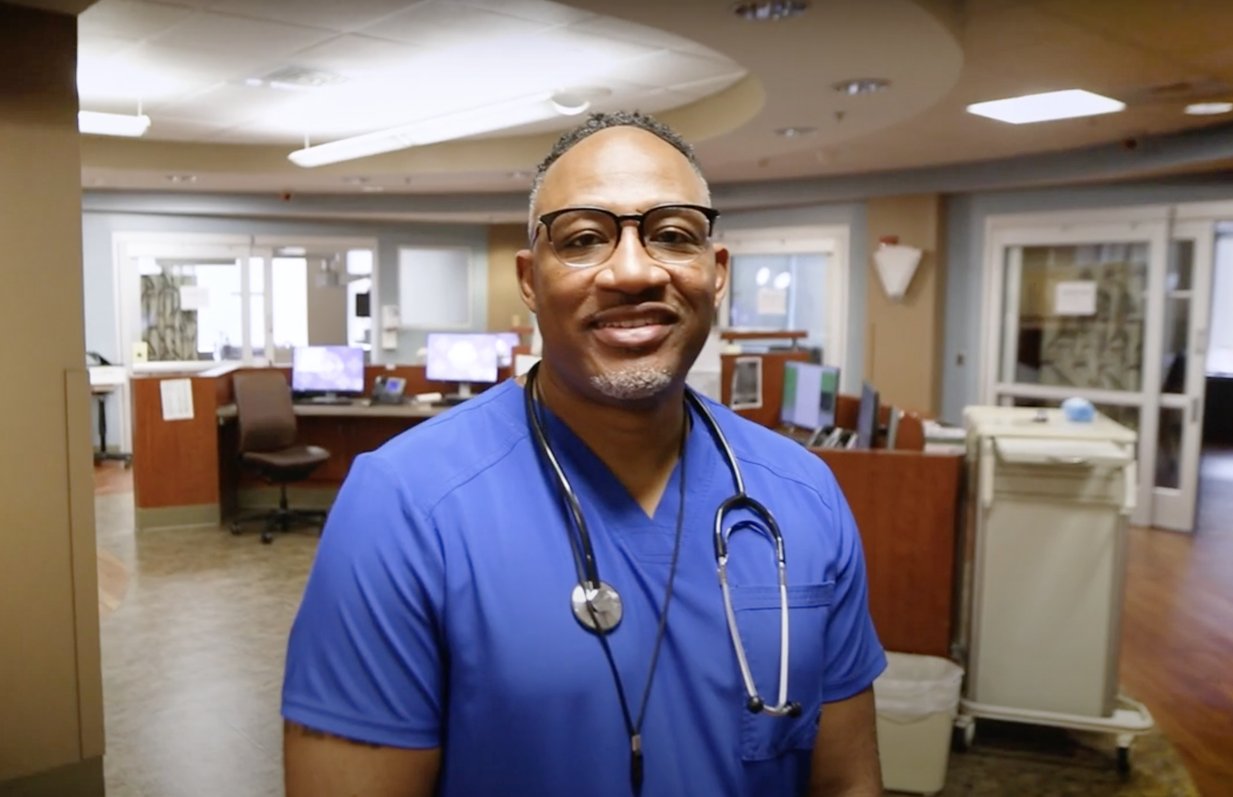A smiling man in black framed glasses and blue scrubs with a stethoscope around his neck.