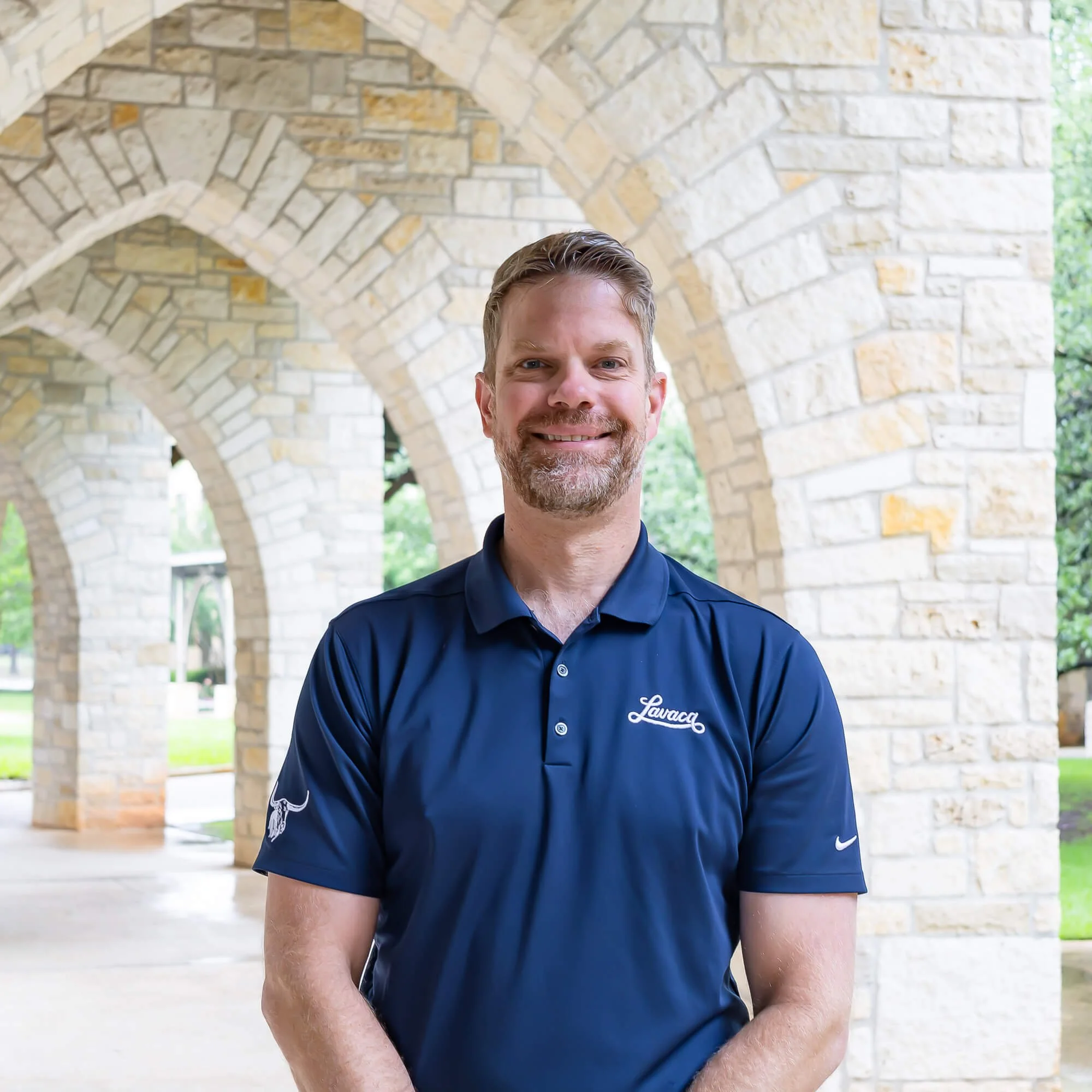 A smiling man wearing a dark blue polo shirt with a logo, standing outdoors in front of a stonearch structure and greenery.
