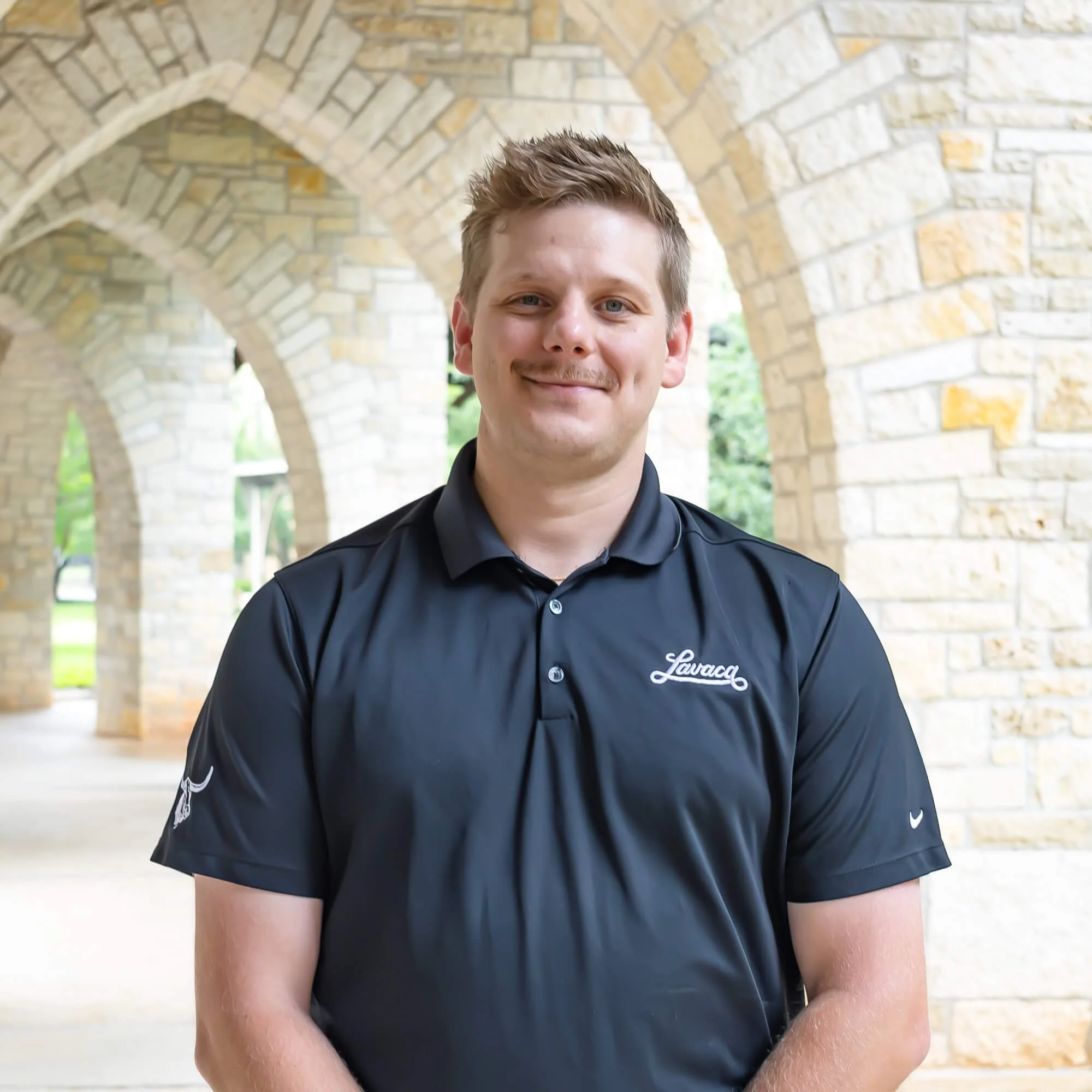 Close-up of a young man with short brown hair and blue eyes smiling, wearing a black collared shirt with a white logo, standing outdoors near a stone archway with greenery in the background.