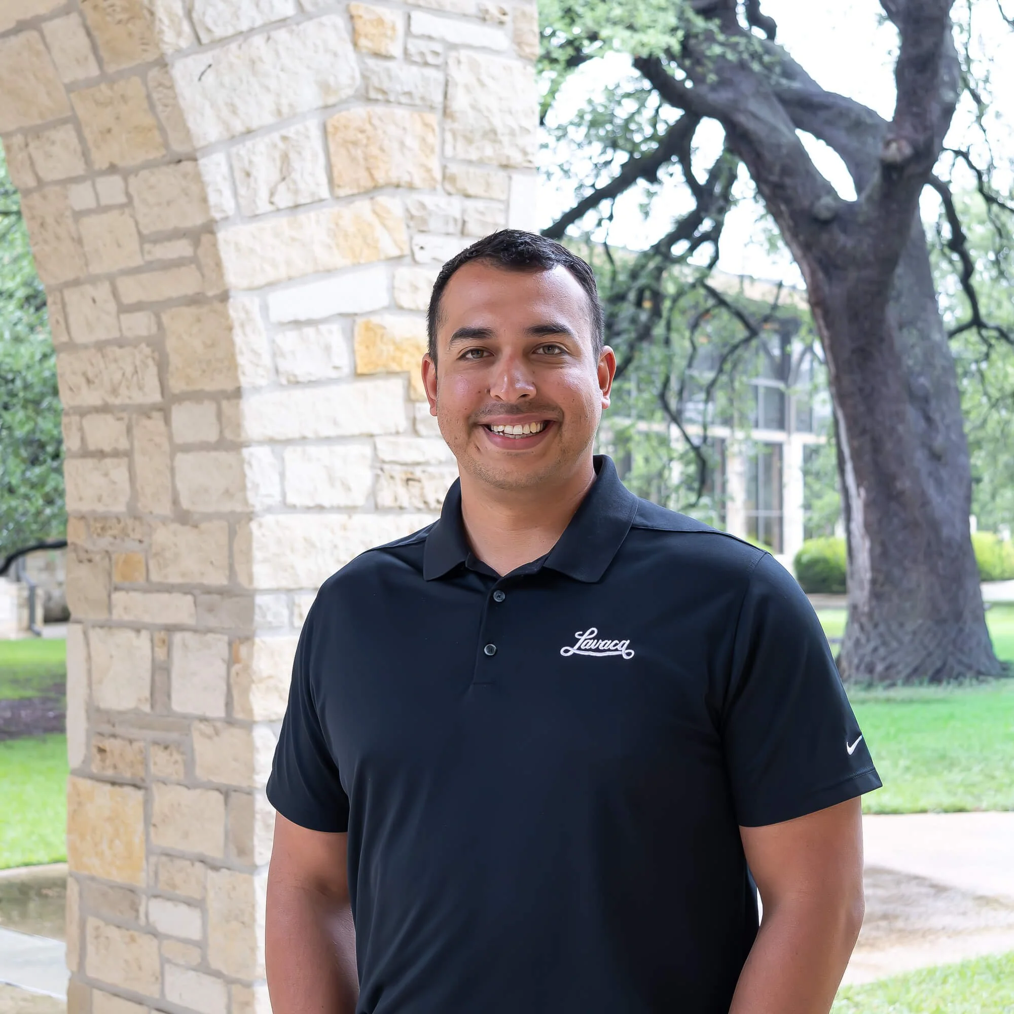 Smiling man in black polo shirt with 'Lavaca' embroidered on it, standing outdoors near a stone building wall and trees.