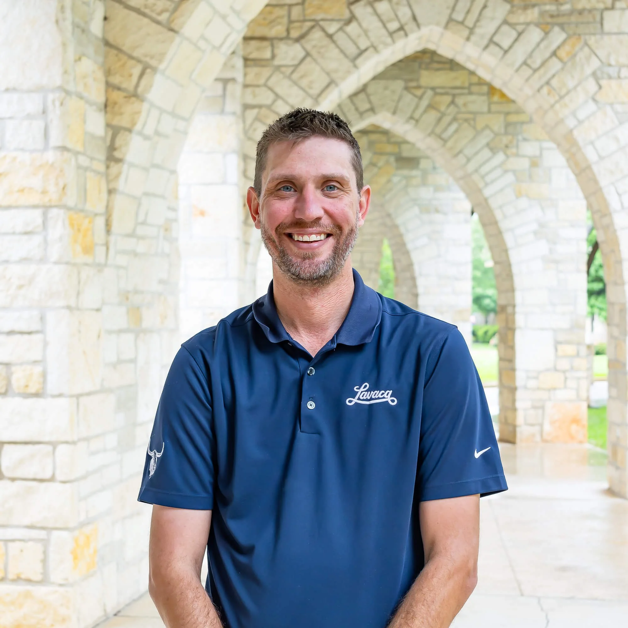 A smiling man with short brown hair and a beard, wearing a navy blue polo shirt, standing outdoors under stone arches with greenery visible in the background.