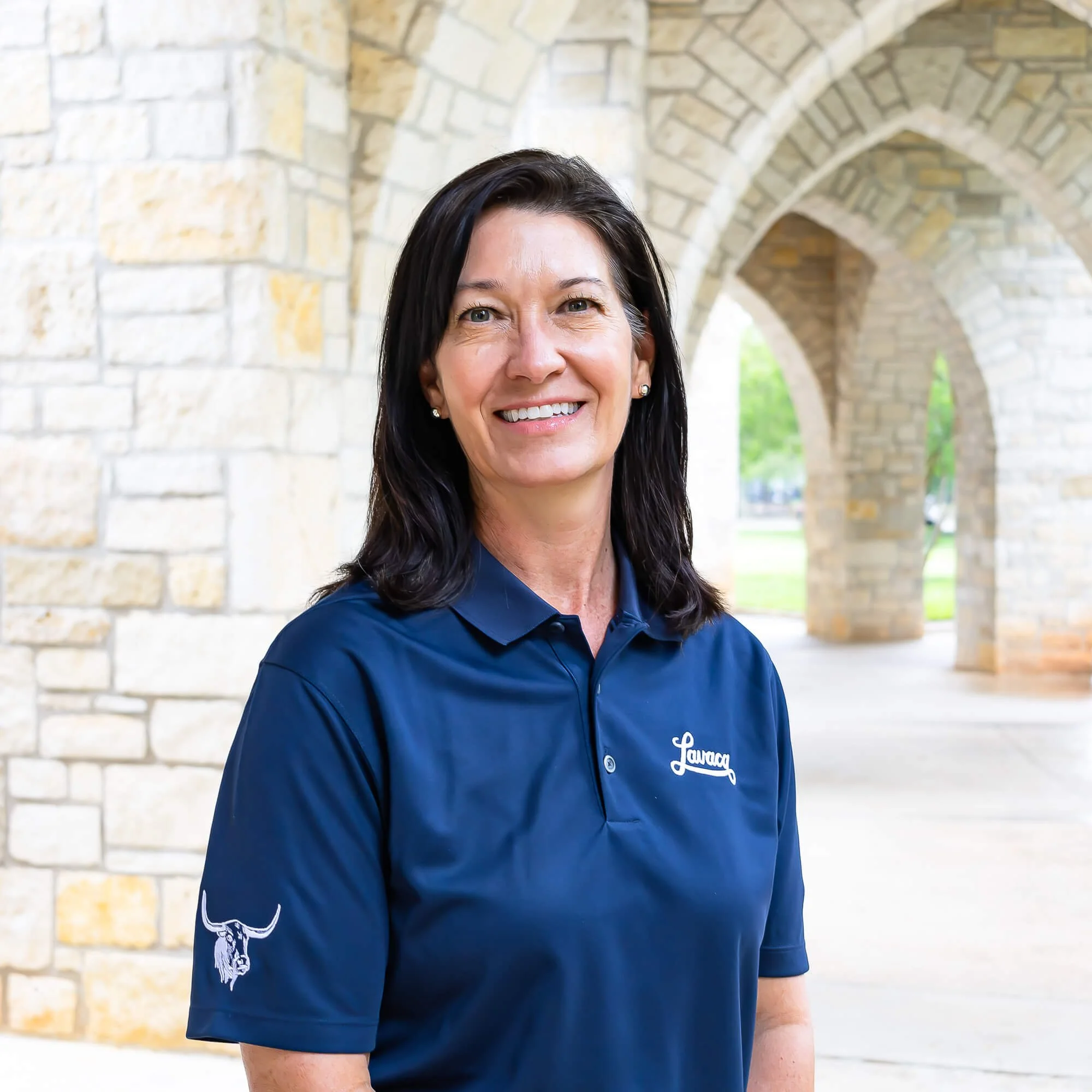 Close-up of a smiling woman with dark hair and earrings, wearing a blue uniform with embroidered logos, standing in front of a stone archway with greenery in the background.
