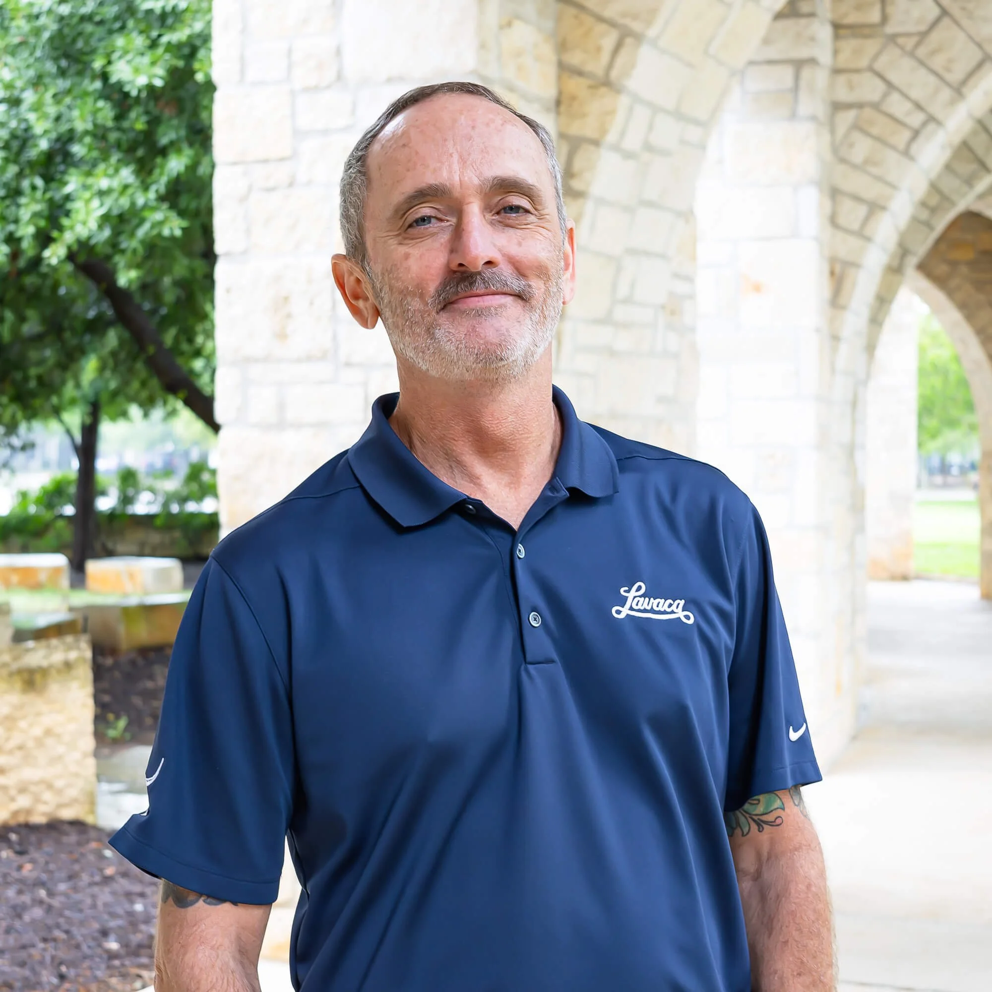 A man with short gray hair and tattoos on his right arm, wearing a navy blue collared shirt with 'Lava' logo, stands outdoors in front of a stone wall and green trees.