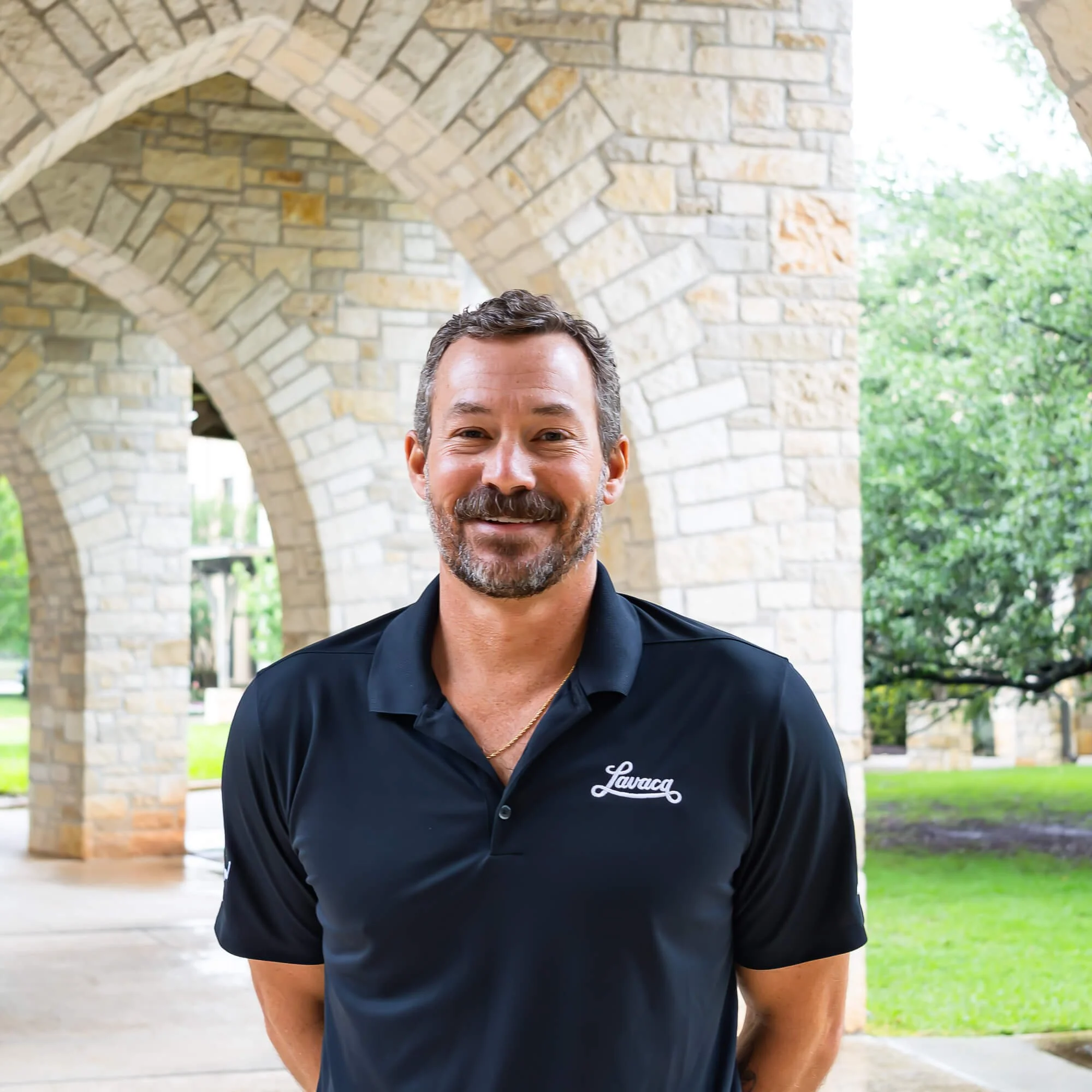Close-up of a man smiling outdoors in front of stone arches and greenery, wearing a dark polo shirt with a logo.