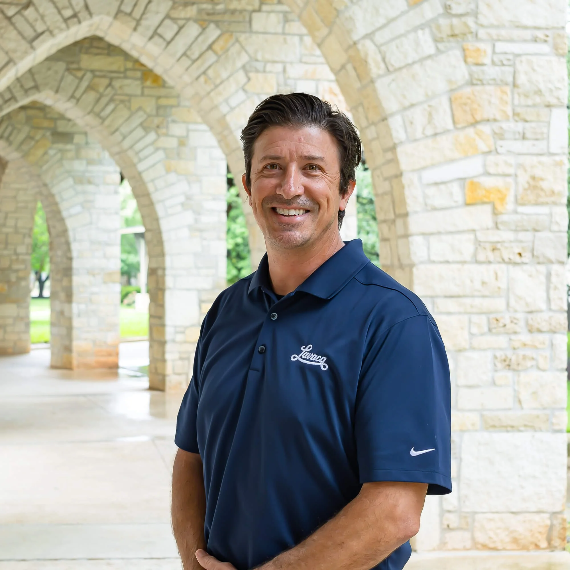 A man smiling wearing a navy blue Nike polo shirt with white writing, standing outdoors near a stone archway with trees in the background.