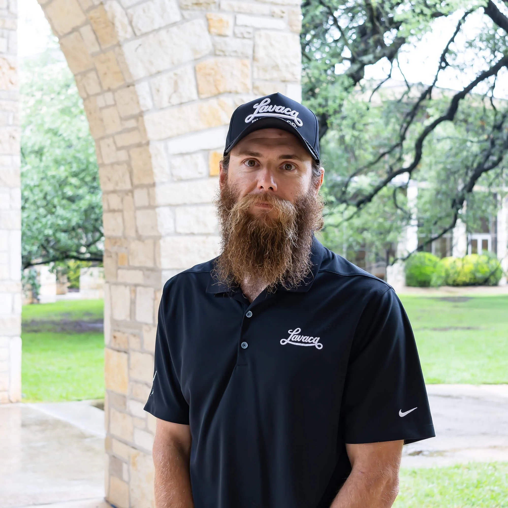 Close-up of a man with a beard wearing a black cap with the word 'Lavage' embroidered on it, standing outdoors near a stone archway and leafy trees.