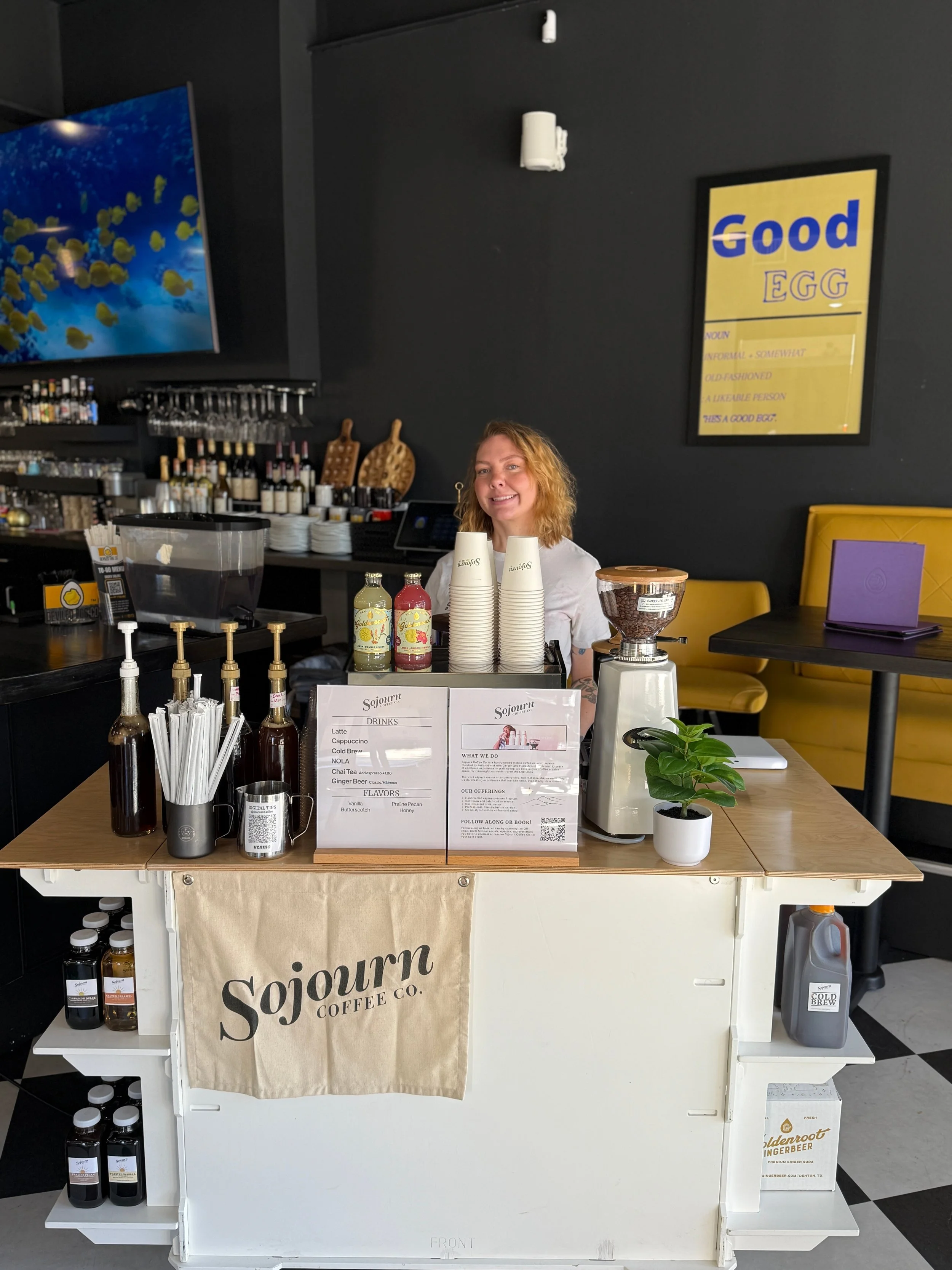 A coffee stand with a woman behind it, featuring bottles of flavored syrups, cups, a coffee grinder, and menu signs, in a modern café setting.