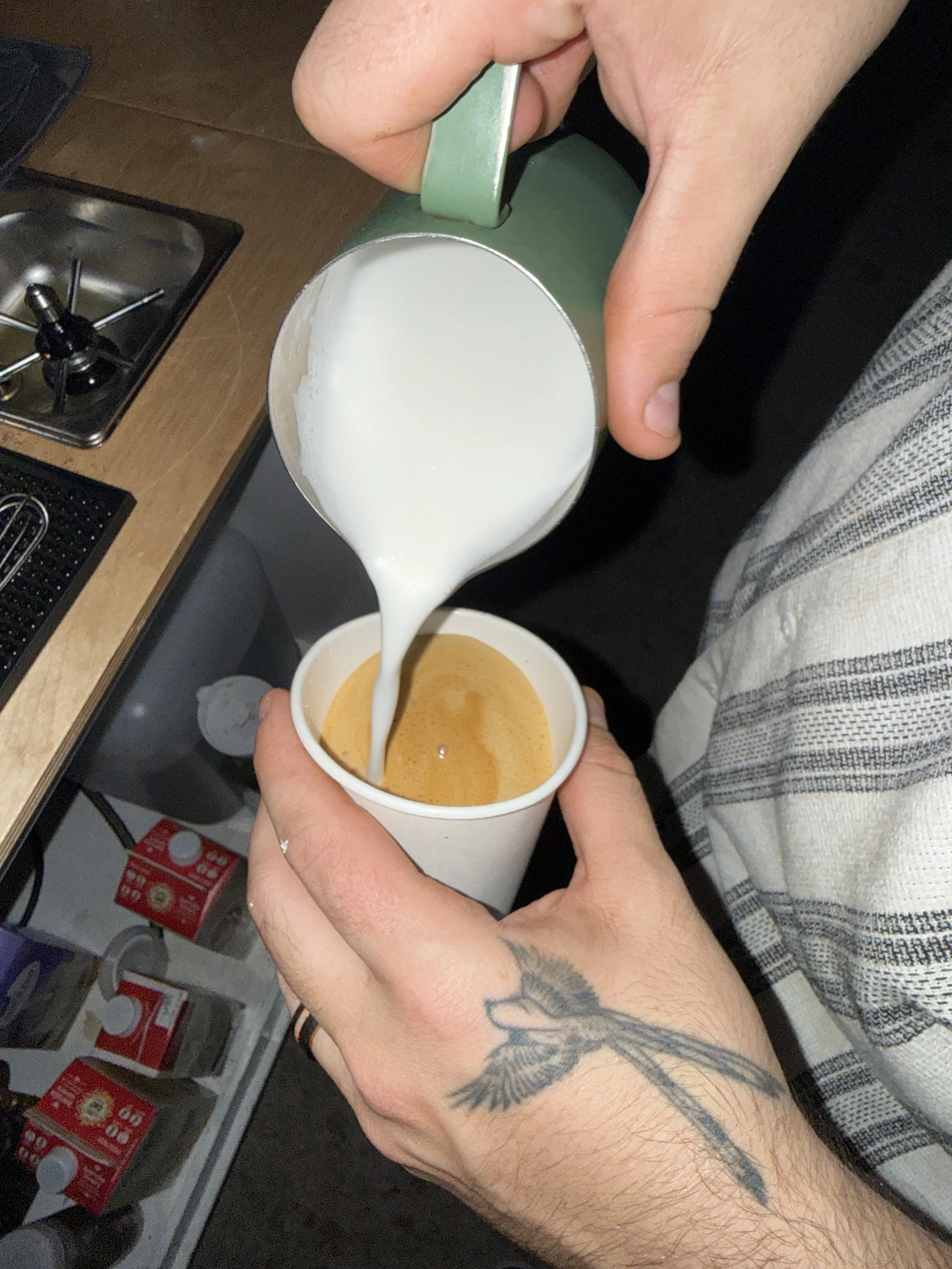 A person with a tattoo on their forearm pouring a light-colored milk or cream into a coffee cup, with store shelves and products visible in the background.