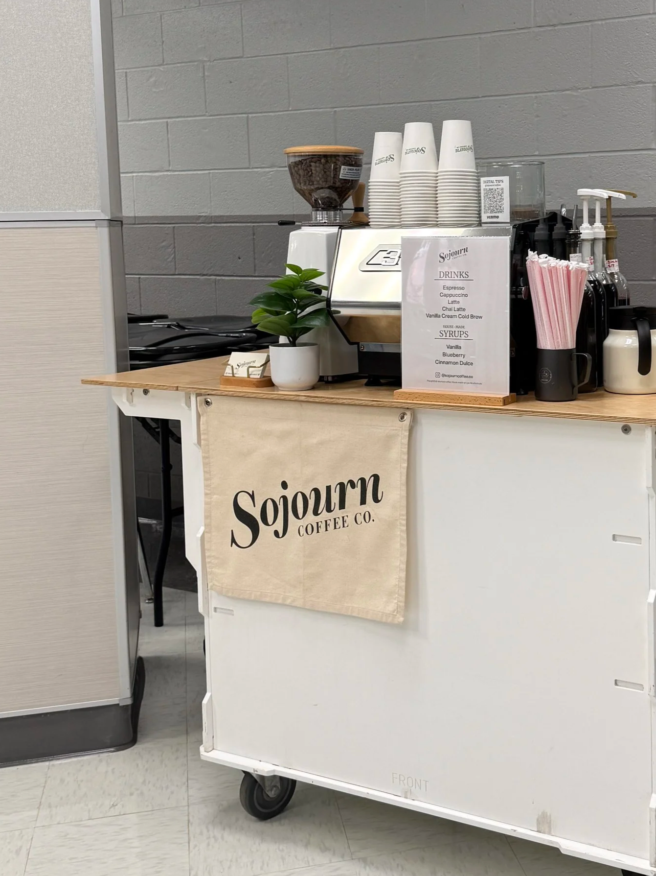 A mobile coffee cart with Sojourn Coffee Co. branding. It has a wooden counter, a small potted plant, a sign with drink and syrup options, and various cups and syrups on top. The cart has wheels.