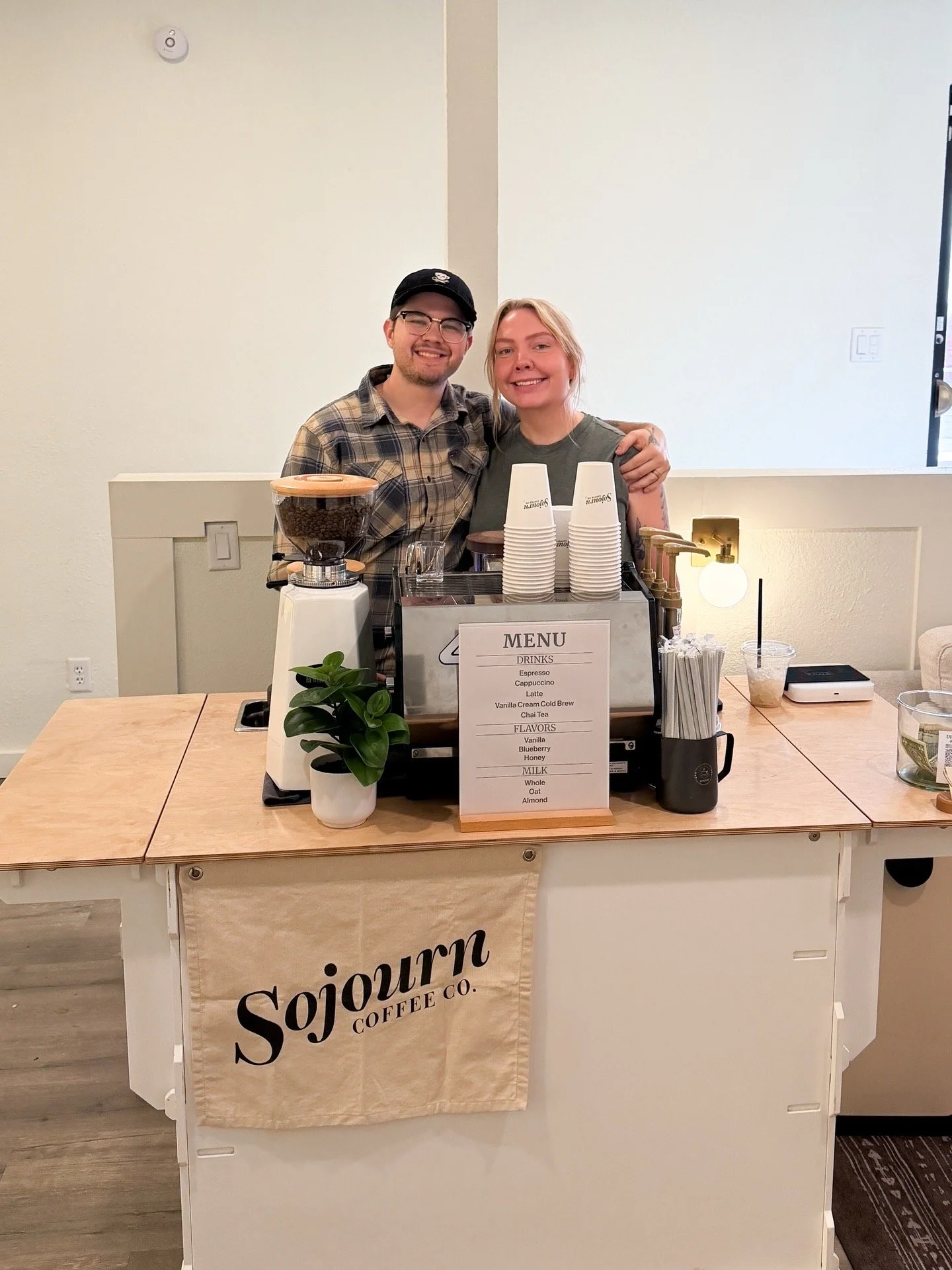 Two smiling people behind a coffee counter at Sojourn Coffee Co., with a menu in front of them, coffee grinder, cups, and a small potted plant on the counter.