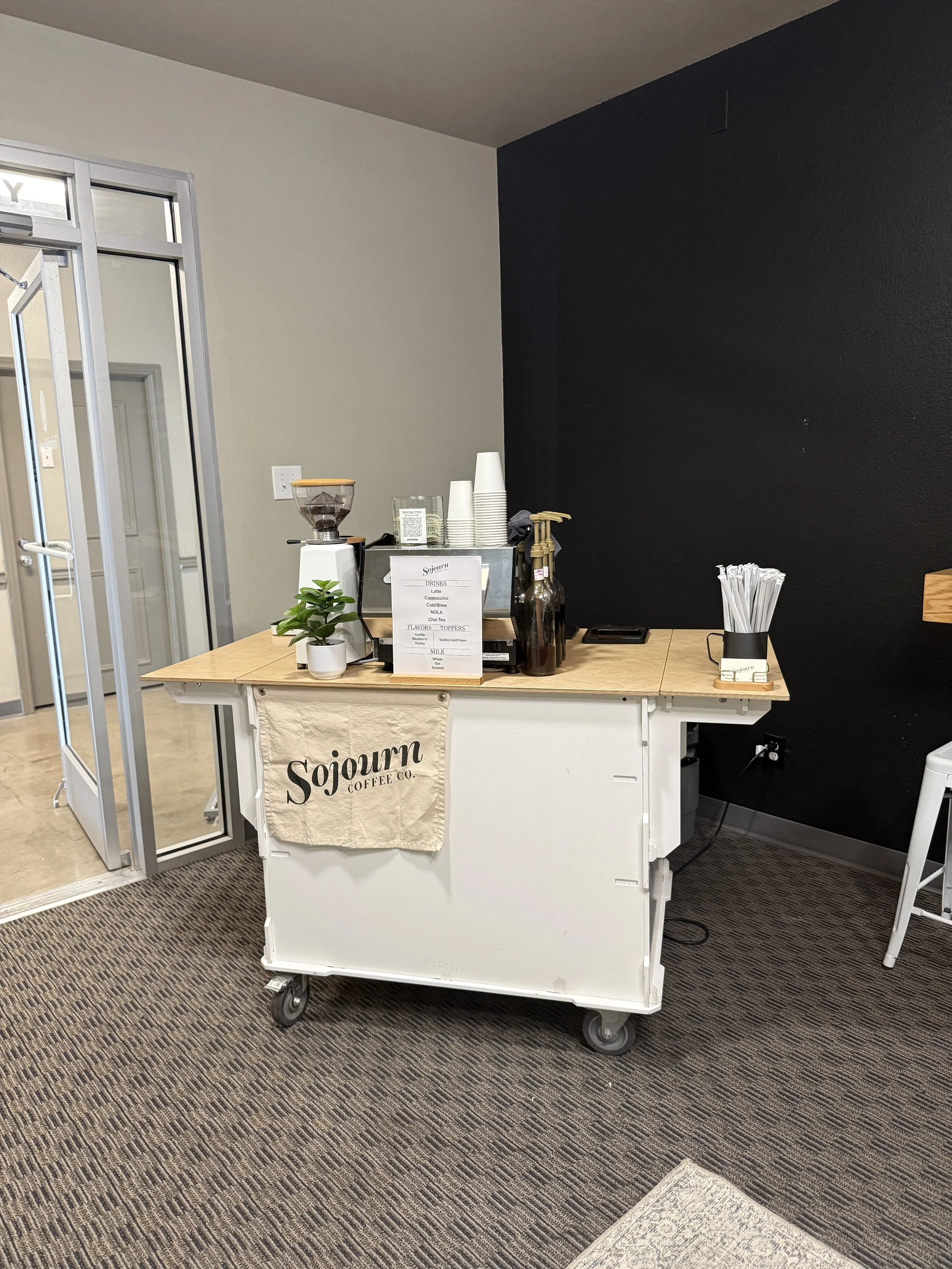 A mobile coffee cart with a black wall background, displaying cups, a coffee grinder, and a menu board for Sojourn Coffee Co., on a patterned carpet floor.