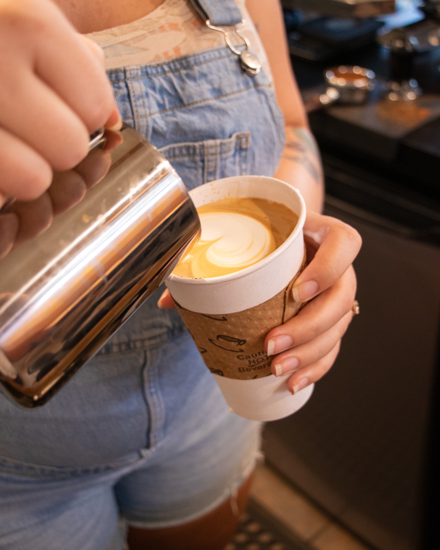 Person wearing denim overalls pouring hot caramel into a takeout coffee cup with a latte art swirl