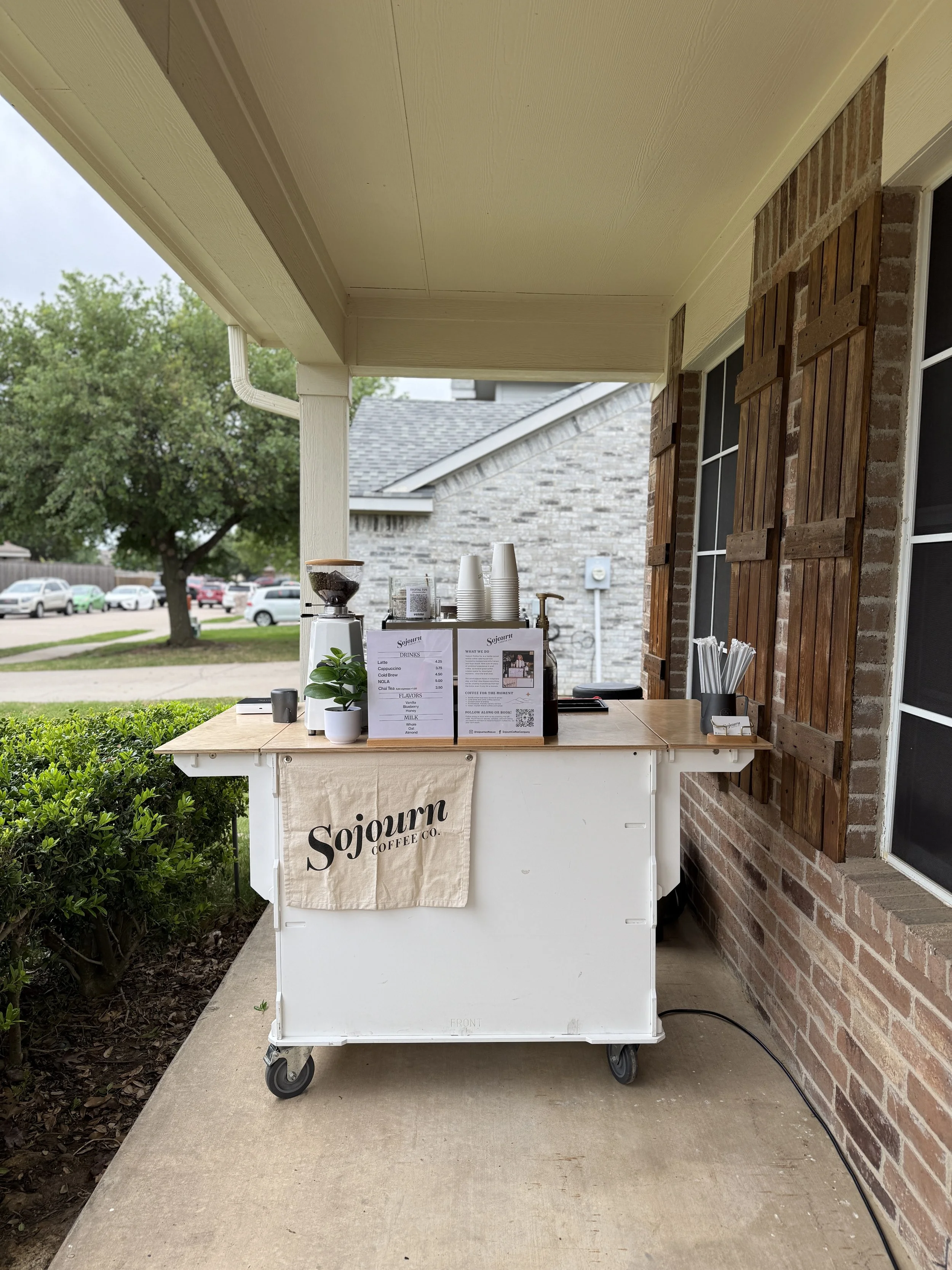 A white mobile coffee cart with a banner that reads 'Sojourn Coffee Co.', set up outside on a sidewalk near a brick building. The cart has a coffee grinder, disposable cups, a small potted plant, and menu signs.