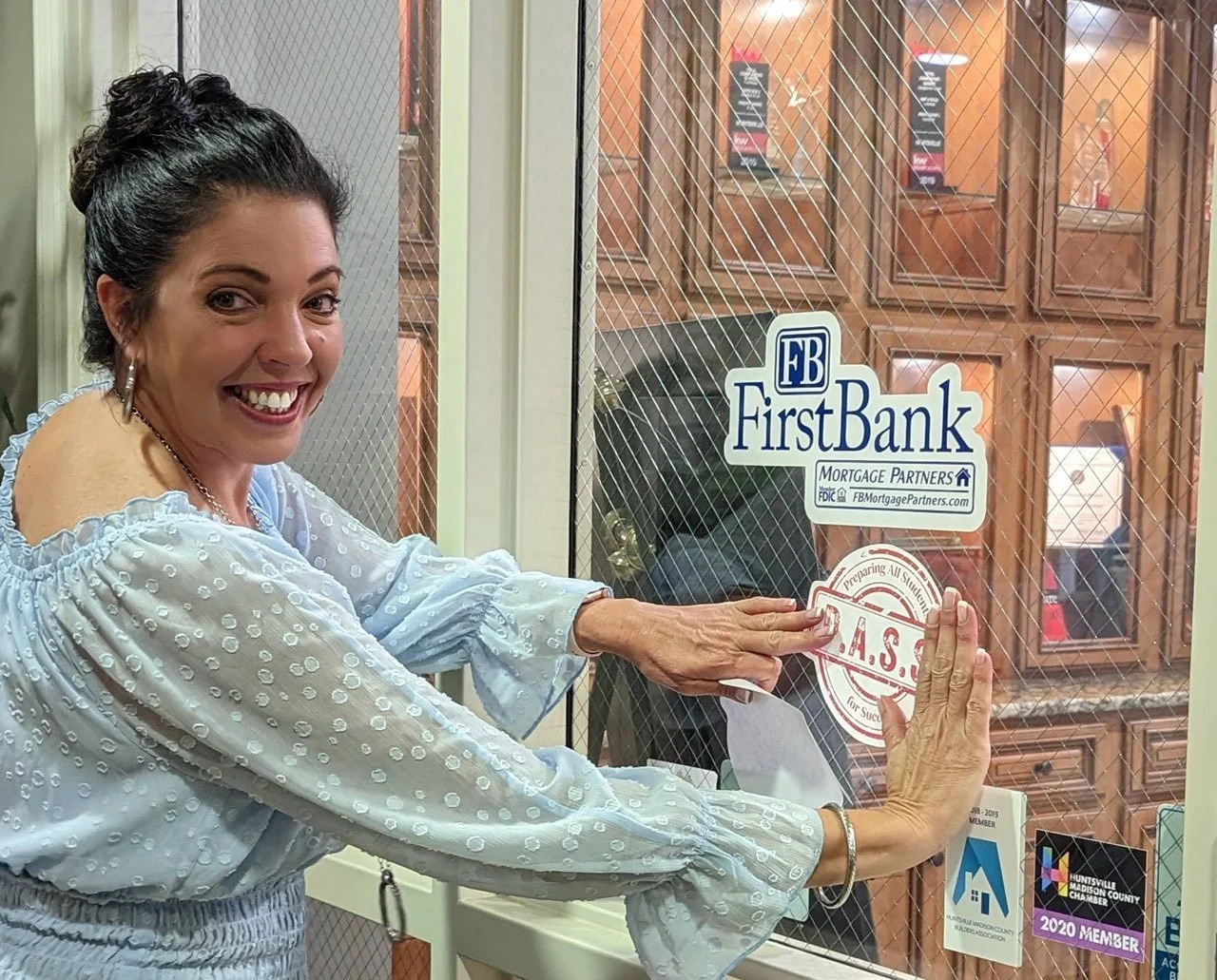 A woman with a big smile, wearing a light blue blouse with polka dots, is placing a sticker on a glass door that has various Business stickers including FirstBank.