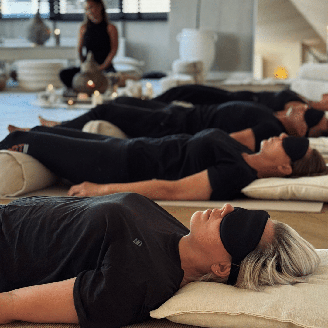 People lying on cushions in a meditation or relaxation session, wearing eye masks, in a cozy indoor setting.