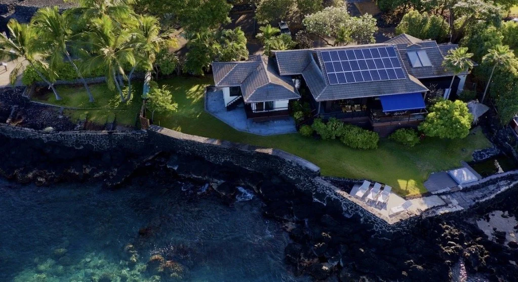 Aerial view of a house on a coastline with palm trees, green lawn, and a rocky shoreline, featuring solar panels on the roof.