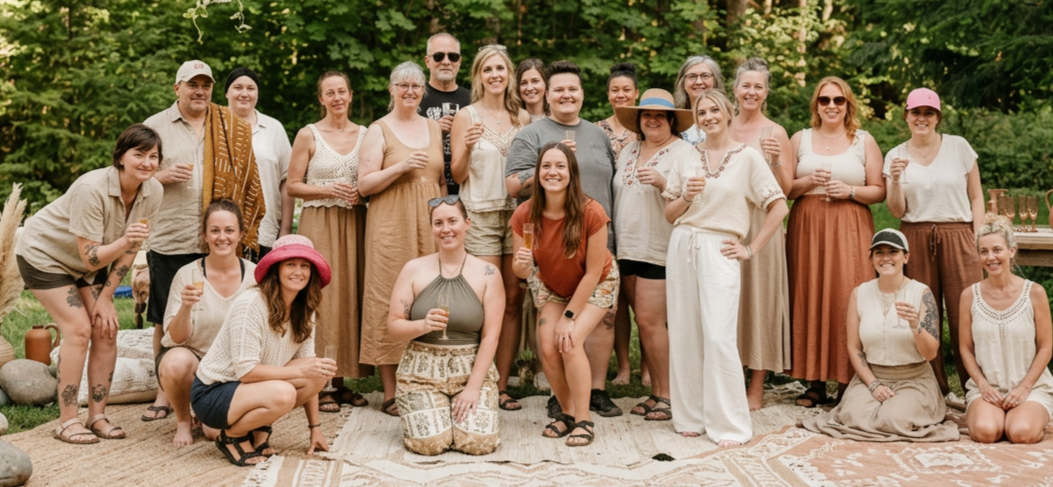 Group of people gathered outdoors, smiling and holding drinks, with trees and greenery in the background, likely at a social event or celebration.