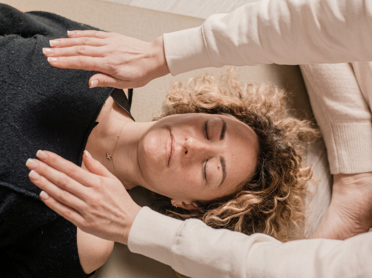 A woman with curly blonde hair lying on her back with eyes closed, receiving a chiropractic or massage adjustment from another person. The second person's hands are placed on her shoulders, and she appears relaxed.