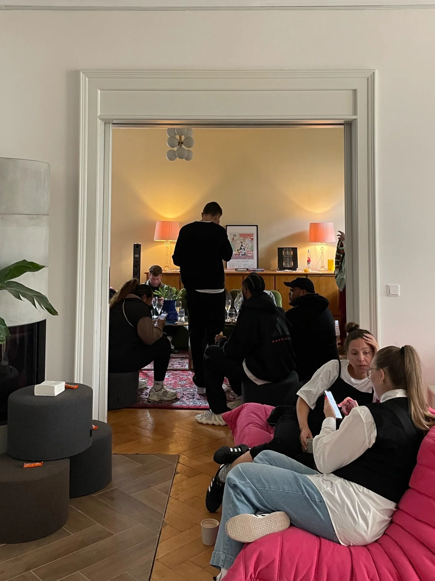 Mehrere Menschen in einem Wohnzimmer, einige sitzen auf einem pinken Sofa, andere am Esstisch im Hintergrund, während ein Mann steht und an einem Tisch arbeitet, vor einem gelben Hintergrund mit Lampen.