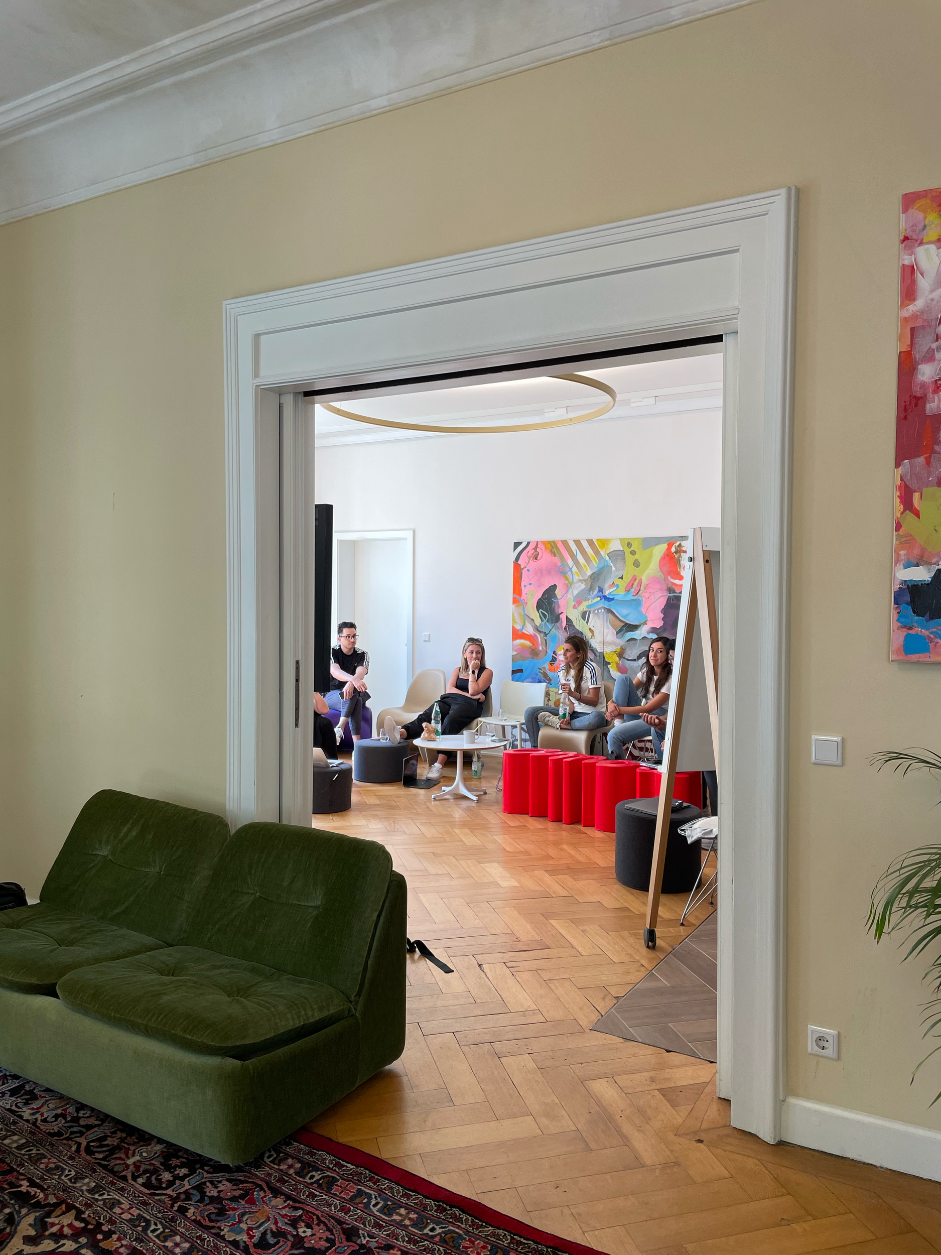 Einblick in einen freundlichen Raum mit Holzfußboden und modernen Möbeln mit mehreren Menschen, die bei einer Diskussion sitzen, bunte Wandkunst im Hintergrund.