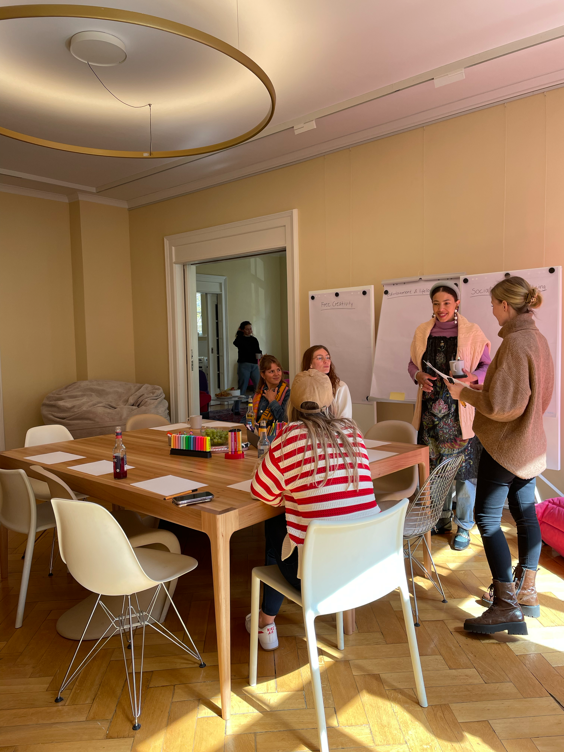 Mehrere Menschen in einem Konferenzraum, die um einen Tisch sitzen und sich unterhalten. Es gibt bunte Marker, Getränke und Snacks auf dem Tisch. Im Hintergrund sind Whiteboards und Flure sichtbar.