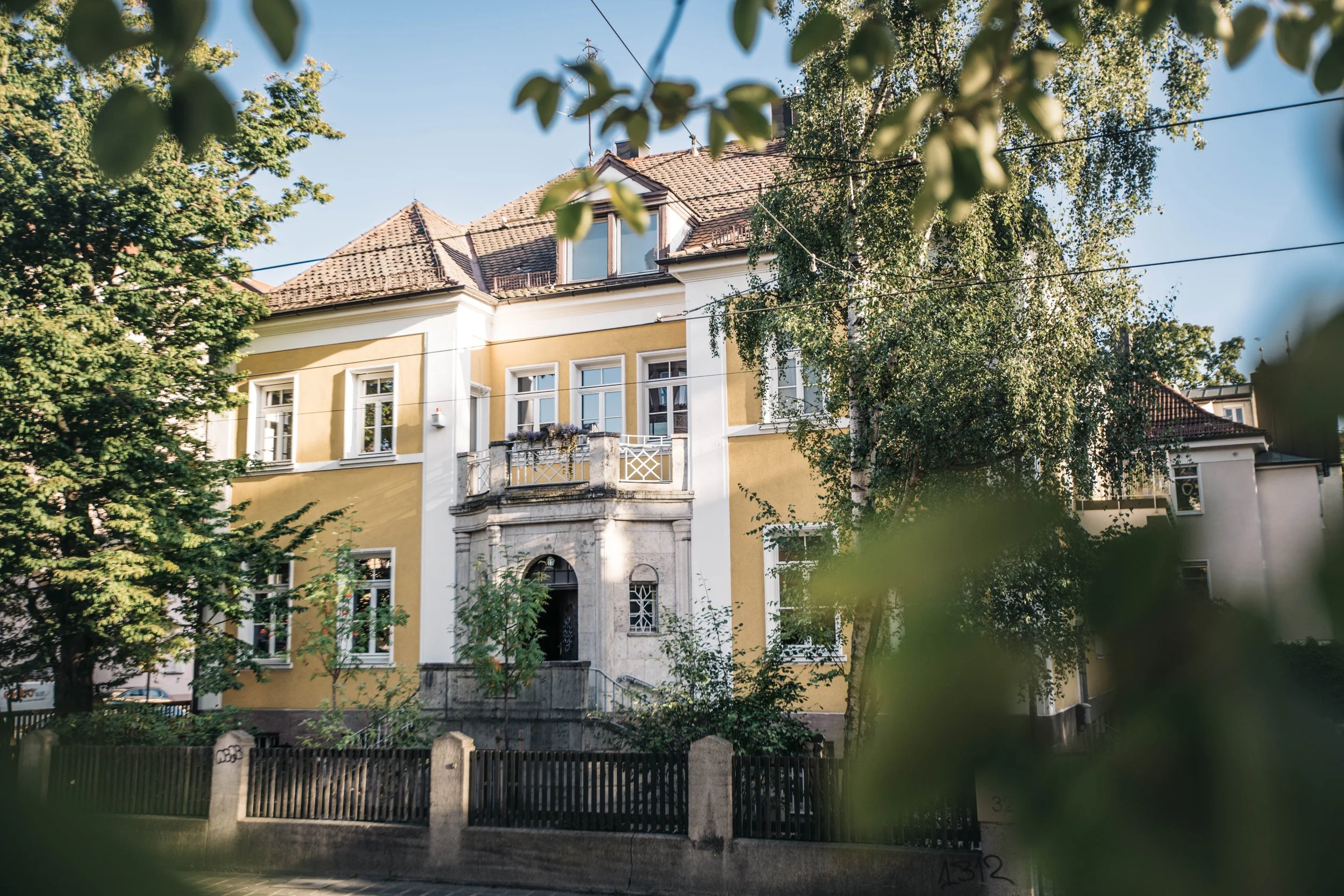 Historisches, mehrstöckiges Haus mit gelber Fassadenelemente, weißen Fensterrahmen und Balkon, umgeben von Bäumen, Blick durch Blätter hindurch bei sonnigem Wetter.