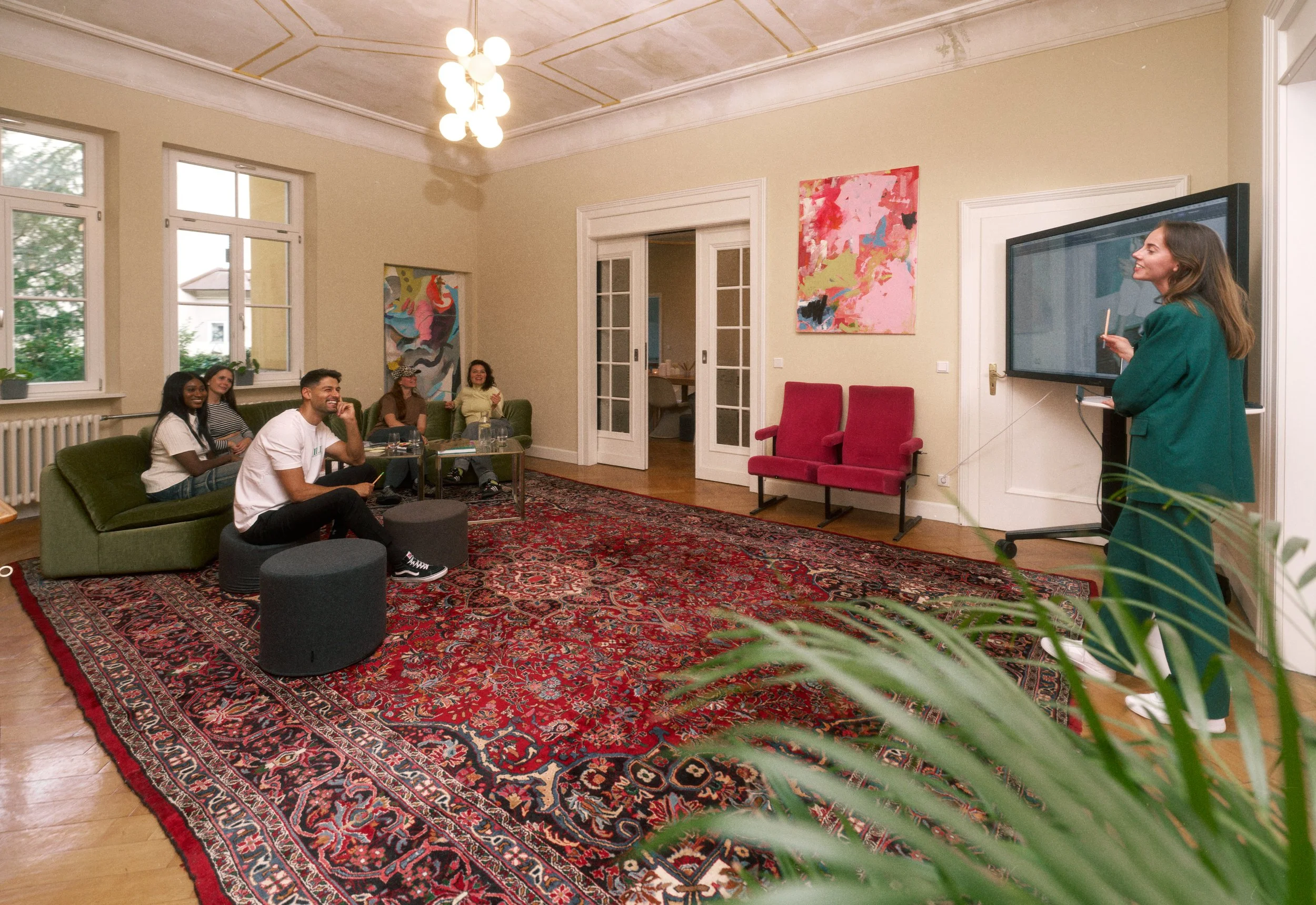 Gruppen von Menschen sitzen im Wohnzimmer und hören einer Frau zu, die an einer digitalen Tafel spricht. Das Zimmer ist hell erleuchtet mit großen Fenstern, bunten Gemälden an den Wänden und einem großen roten Teppich auf dem Holzfußboden.