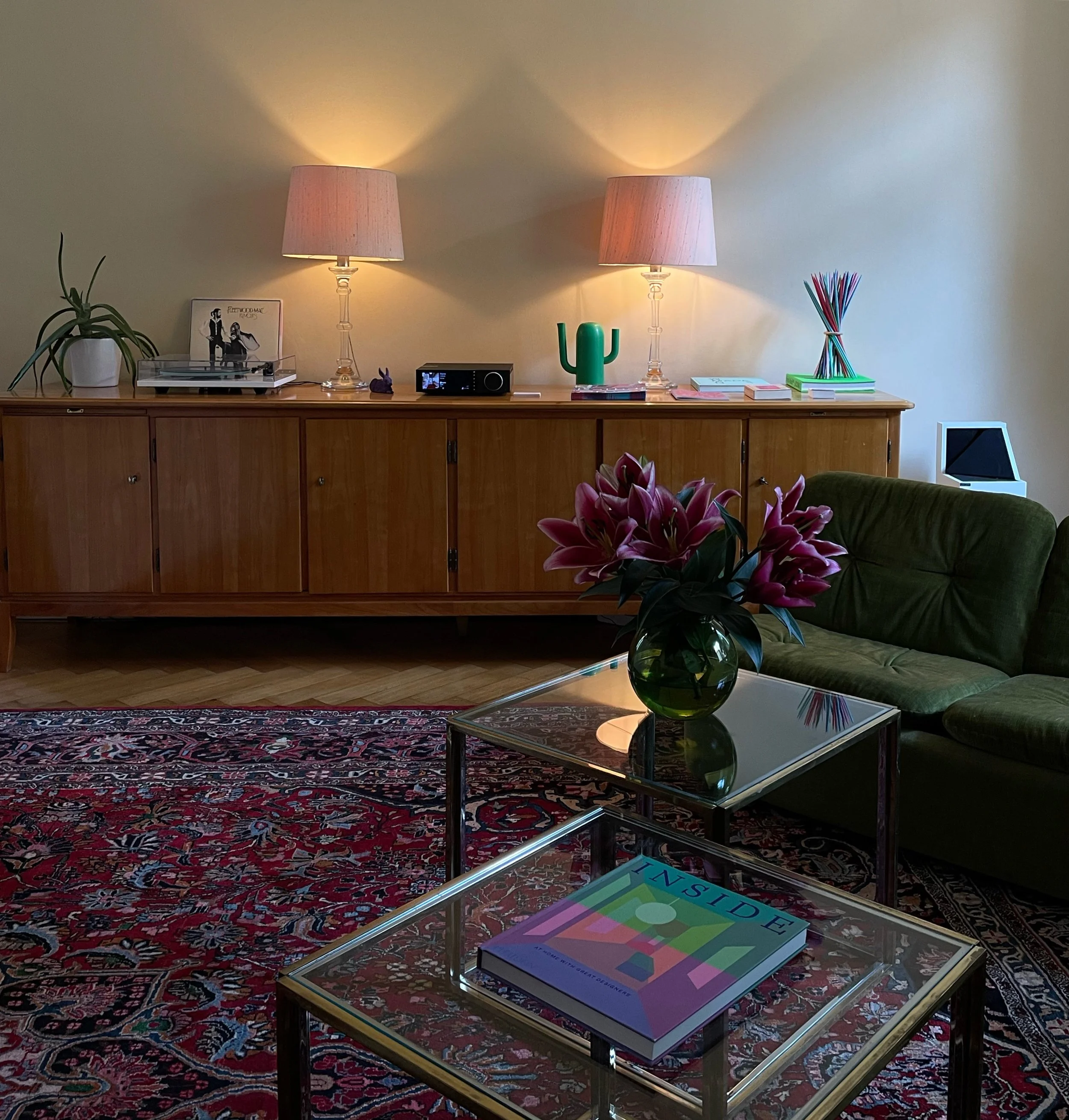 Gemütliches Wohnzimmer mit grünem Sofa, Tisch mit Blumen, Holzsideboard mit Lampen, Pflanzen und Dekorationsgegenständen, Teppich, Tablet auf einem Ständer, Buch auf Couchtisch