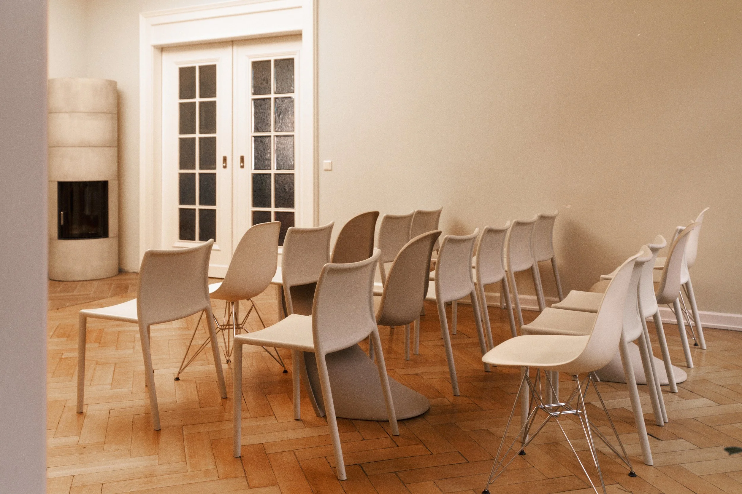Leerer Raum mit acht weißen Stühlen auf Holzboden, eine Wand mit Doppelglastür, eine Ecke mit Kamin.