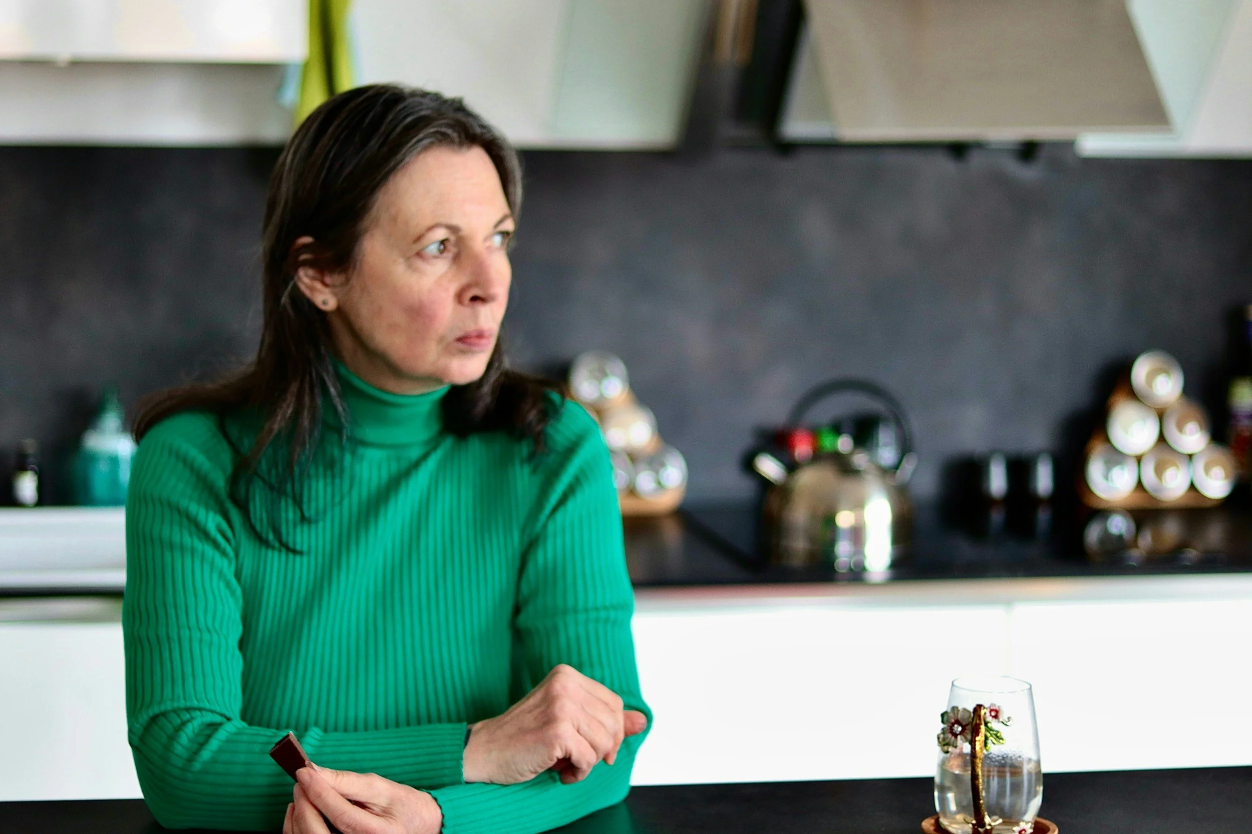 A woman with dark hair in a green turtleneck sitting at a kitchen counter with a glass of water and decorative necklace in front of her, looking to her left.