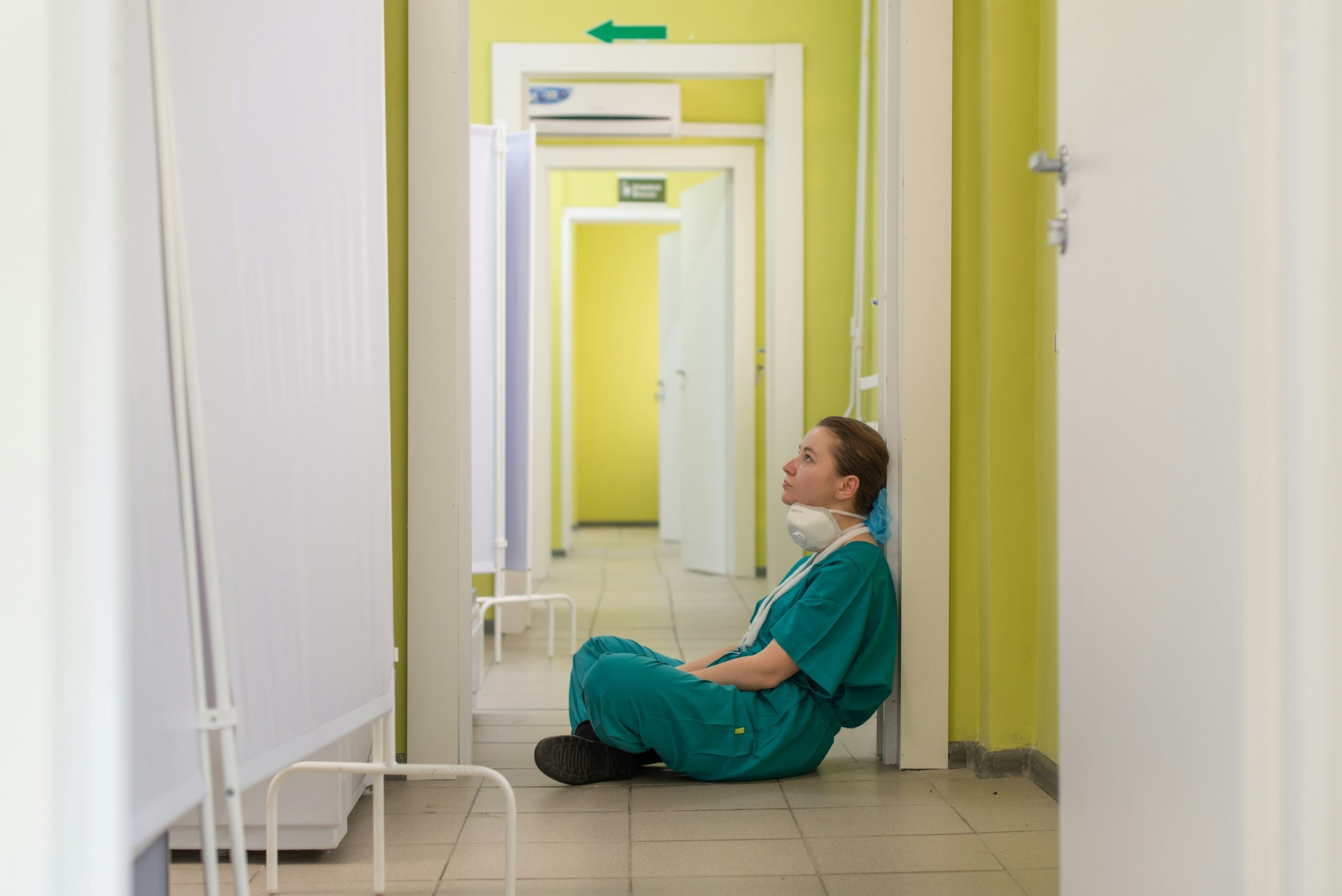 A healthcare worker in scrubs and a face mask sitting on the floor in a hospital corridor.