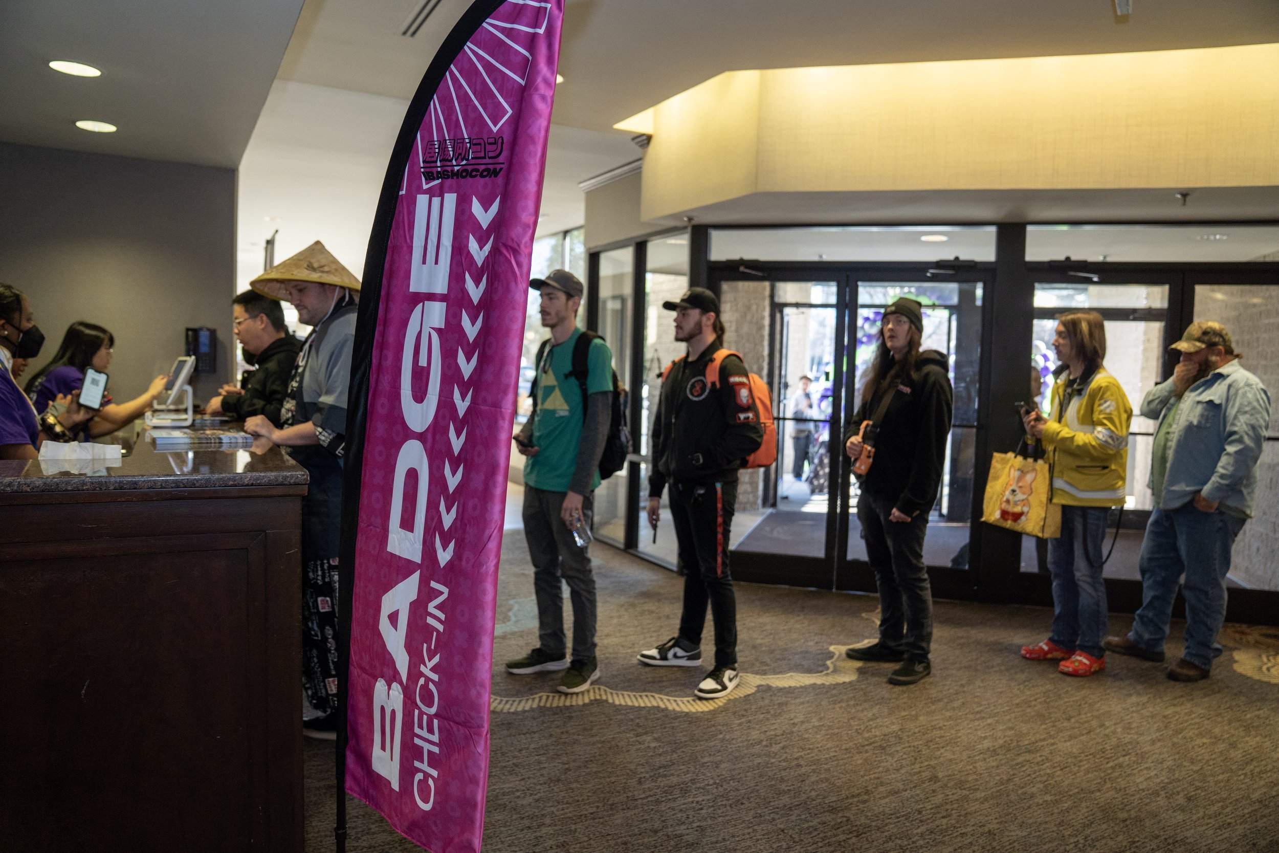 Group of people standing in line at the check-in desk of a convention center, with some looking at their phones, and a pink banner displaying the event name, BASHOCON, in front of the desk.