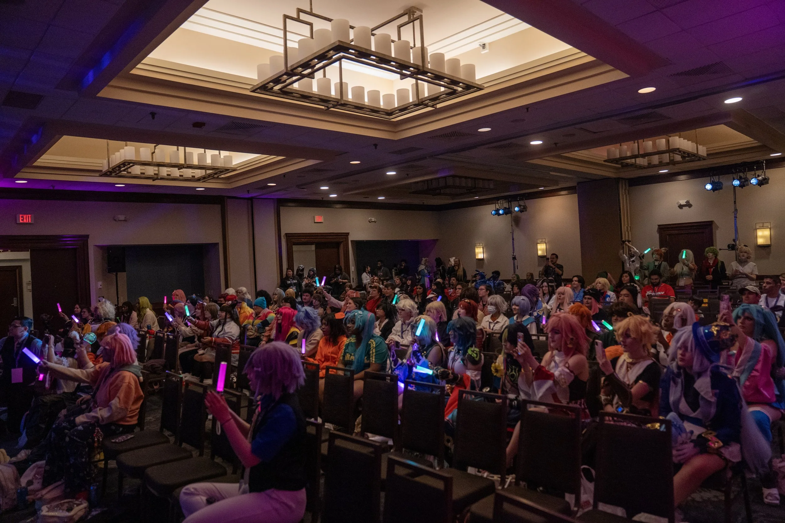 A large group of people cosplaying as anime characters sitting in an auditorium, holding glowing light sticks during a convention or cosplay event. This was the Idol fest event at IbashoCon 2025.