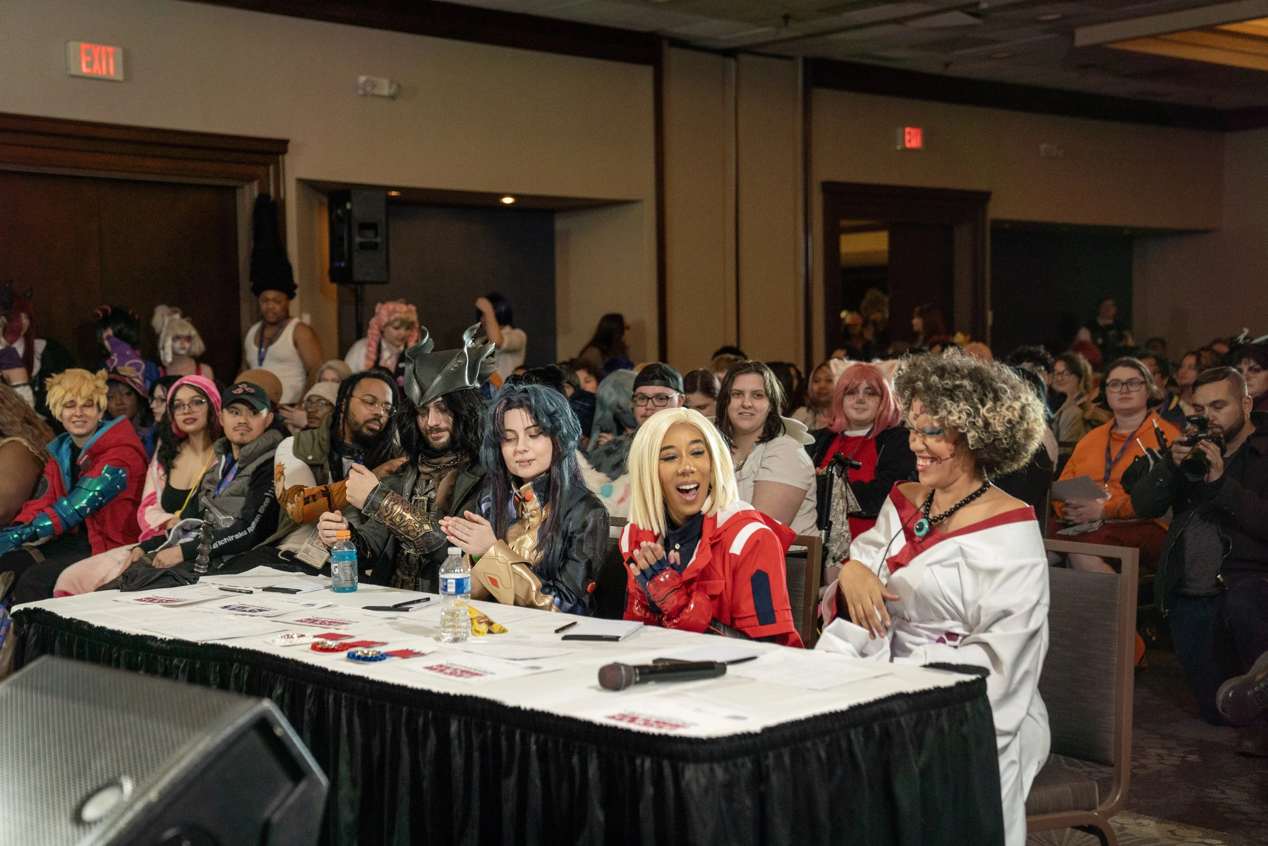 A large group of people dressed in cosplay at IbashoCon, with some judges sitting at a long table and an audience sitting behind them.