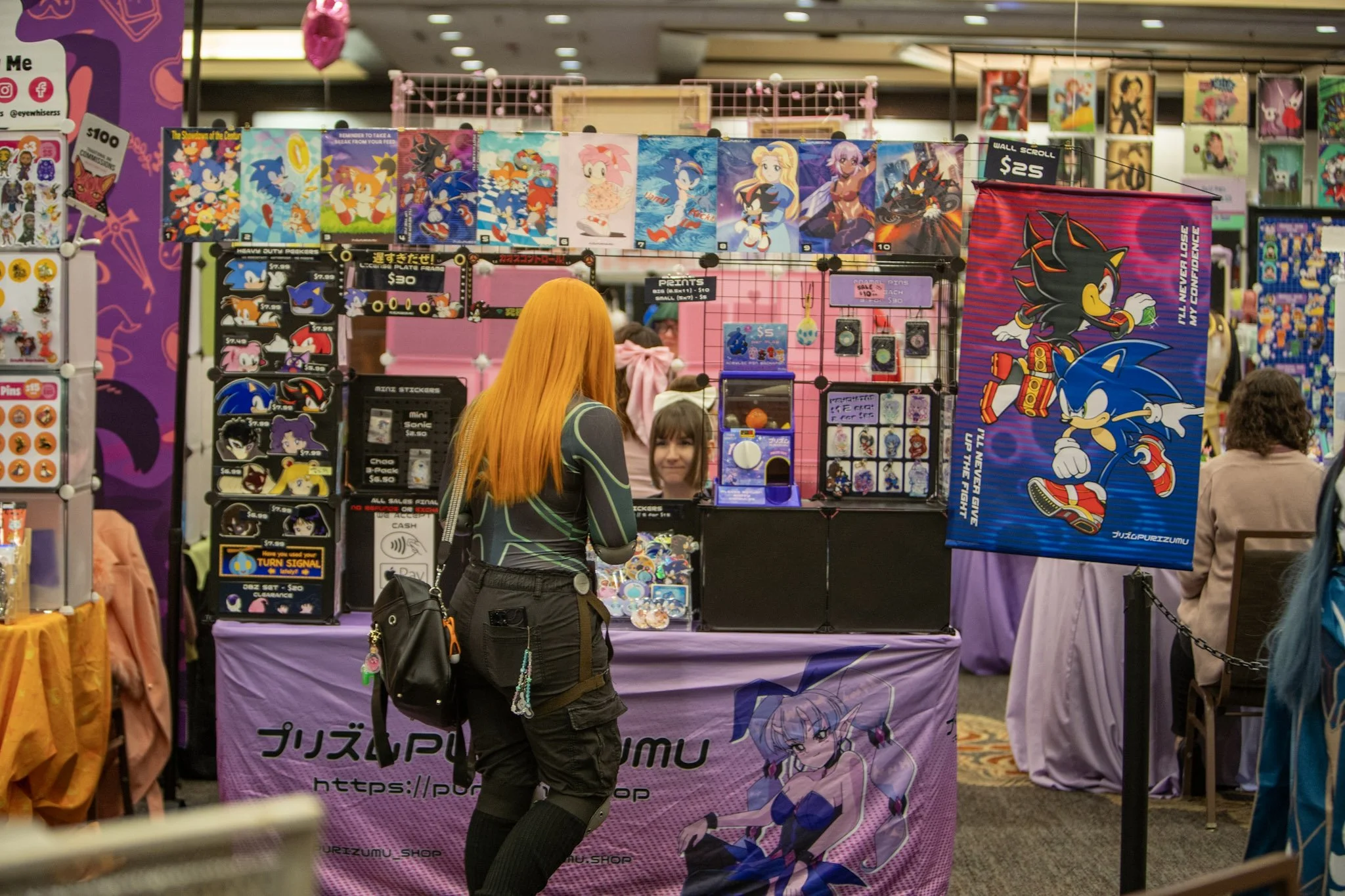 A woman with long orange hair browsing a vendor stall at IbashoCon. The stall features Sonic the Hedgehog merchandise, including stickers, buttons, and posters. There is a large Sonic poster and various merchandise displays.