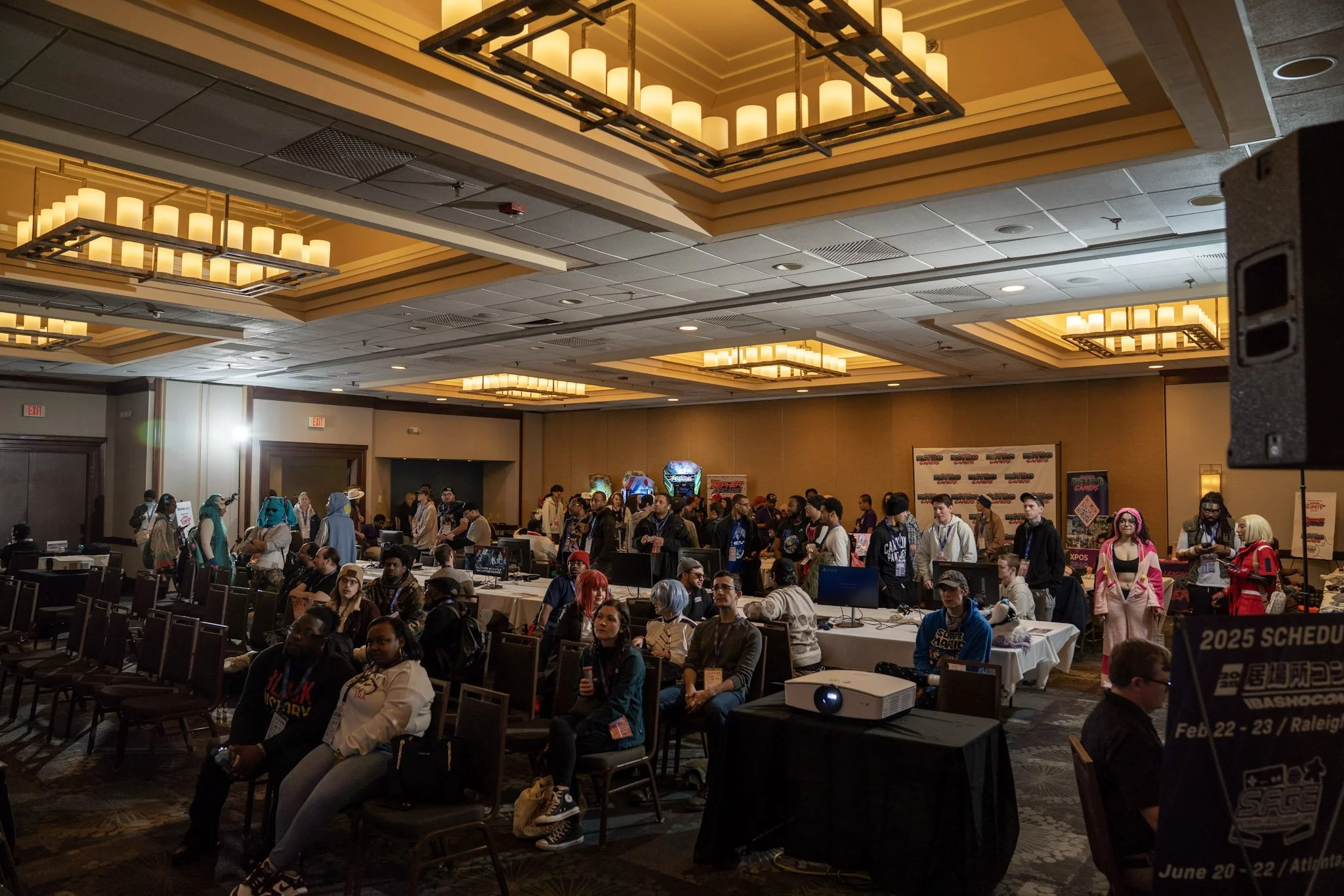 A crowded ball room  with numerous attendees, some seated and some standing, many wearing cosplay costumes, and arcade machines in the background. IbashoCon 2025 game room for free play and Ibashowdown (Formerly known as Gohon Kumite)