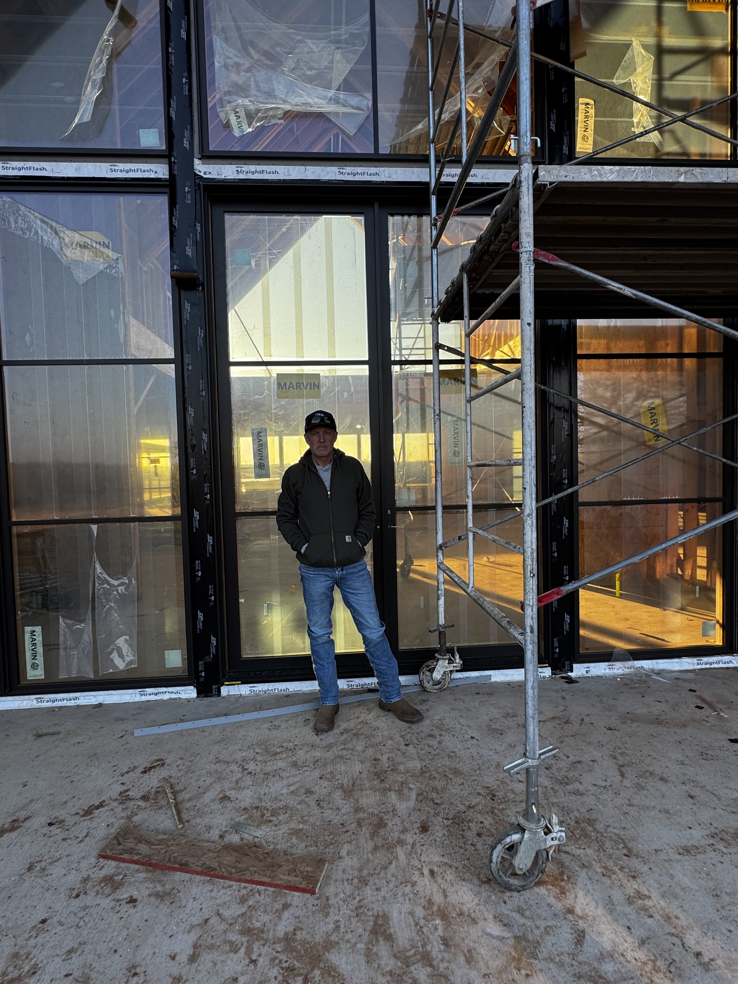 Shon stands in front of the newly installed front doors at The Essex Ranch, marking a major step forward in the build. The sleek black-framed glass entry, now in place, reflects the golden light of a day’s work well done—one more vision turned into r