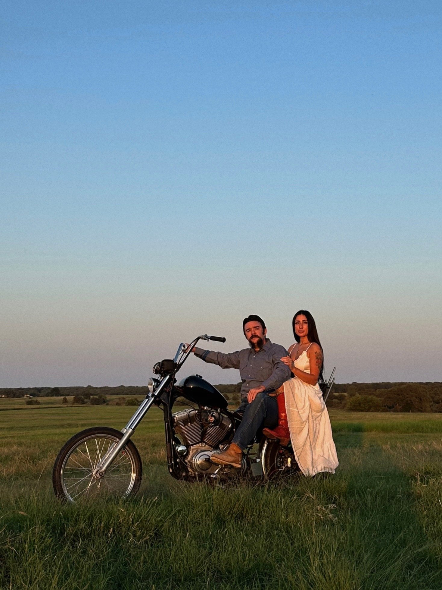 A beautiful photo of Hannah and Tanner on Tanner’s custom motorcycle, set against the backdrop of the coastal hayfield during golden hour. The soft golden light envelops them, creating a romantic atmosphere as Hunter captures their engagement photos,