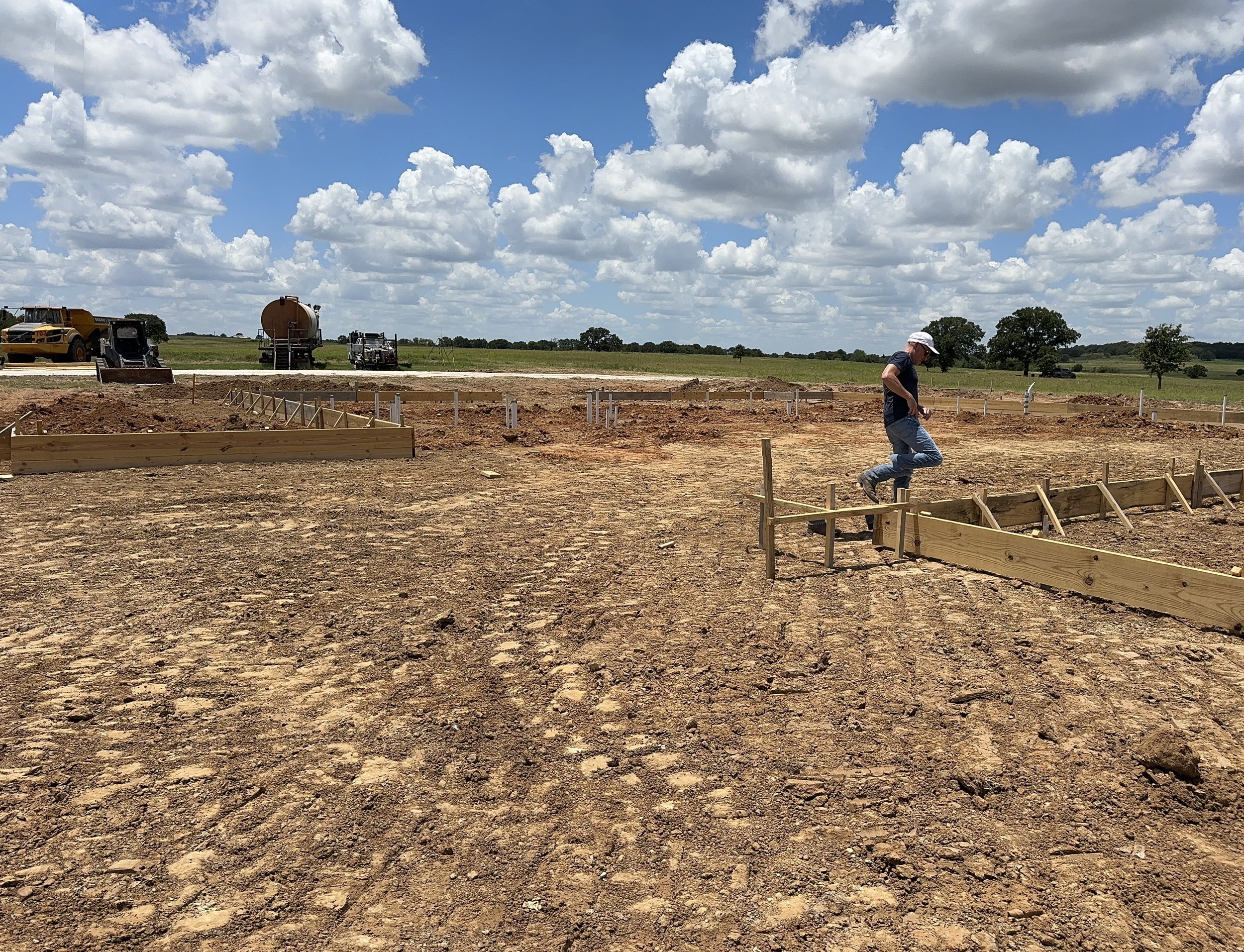 A beautiful day at The Essex Ranch with blue skies and pillow-like clouds overhead. The site has been graded, the forms are set, and the electrical and plumbing are being installed in the background. Shon inspecting the progress, as the foundation of
