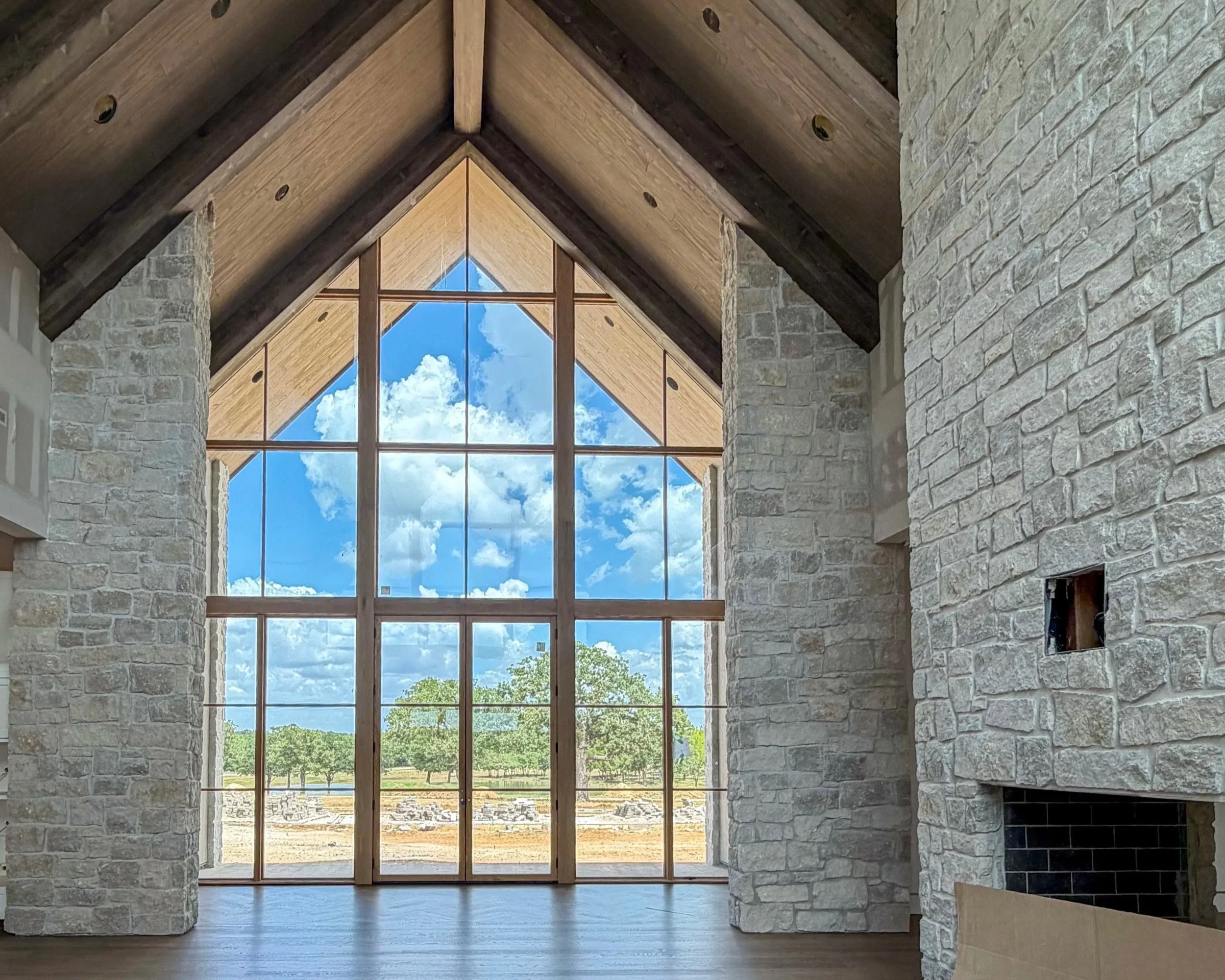 Great room at The Essex Ranch with a vaulted wood ceiling, floor-to-ceiling bronze-framed windows, pale stone walls, and views of the Texas landscape.