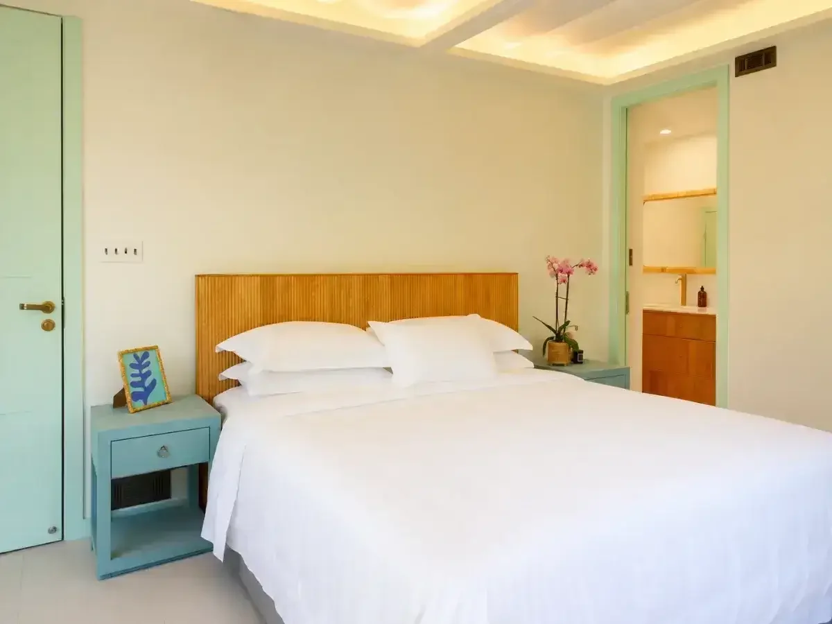 A white bed with multiple pillows in a bedroom with pastel-colored decor and a built-in niche with shelves and a small cabinet.