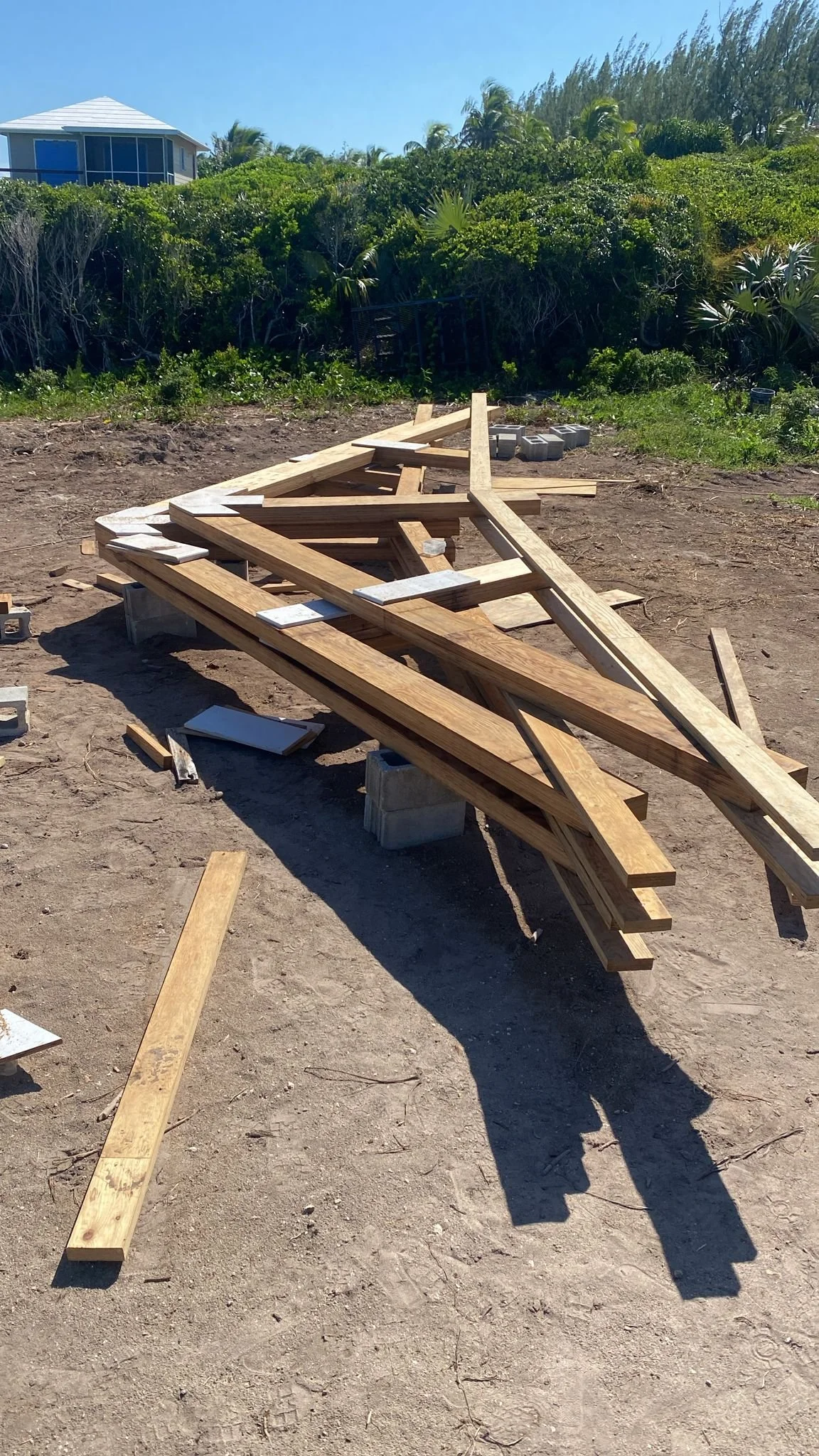 Construction site with wooden beams and concrete blocks on dirt ground, with green bushes and a house with a blue roof in the background under a clear blue sky.