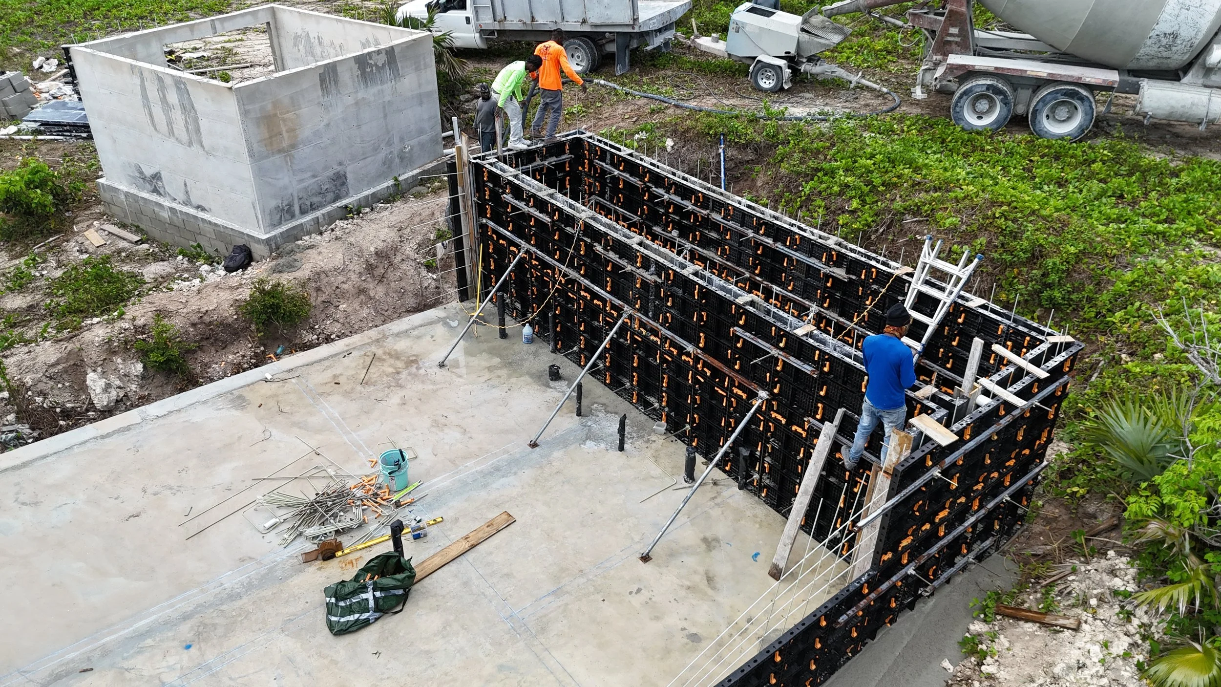 Construction workers are building a foundation with formwork and rebar, with a concrete mixer truck and construction equipment in the background.