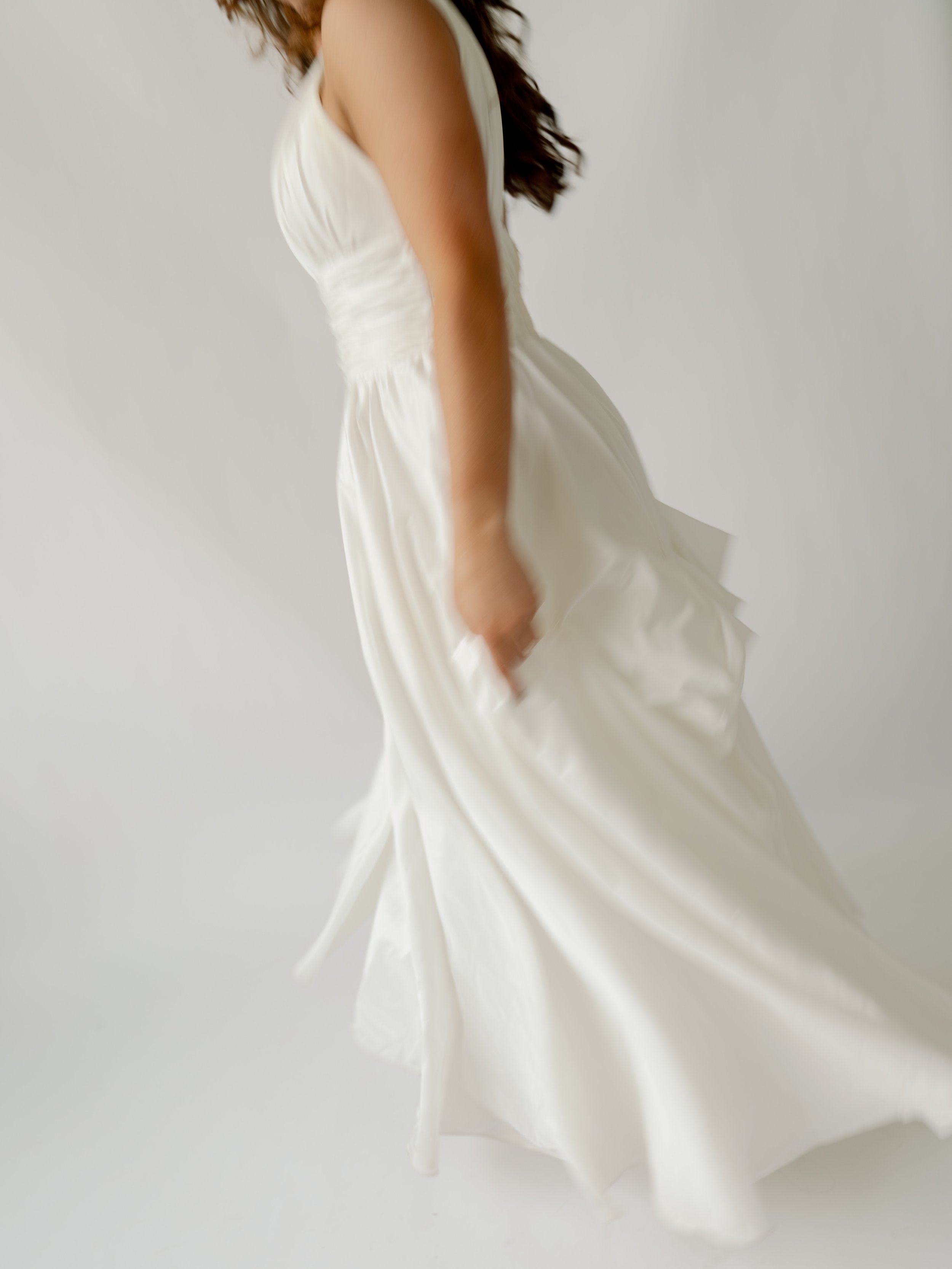 Close-up of a woman in a white wedding dress, holding the skirt with her right hand, against a plain background.