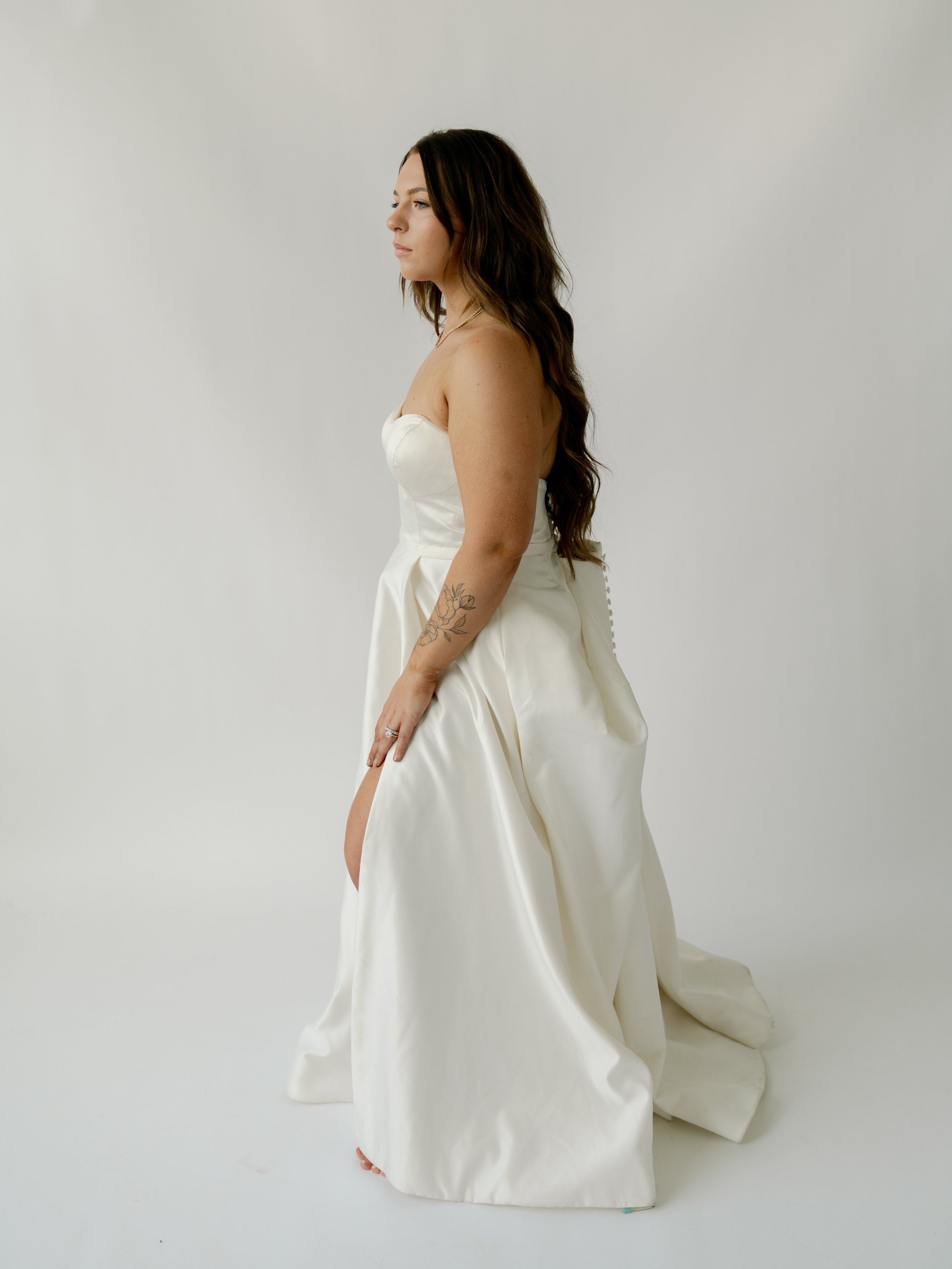 A woman in a strapless white wedding gown with a slit in the skirt, standing sideways against a plain white background.