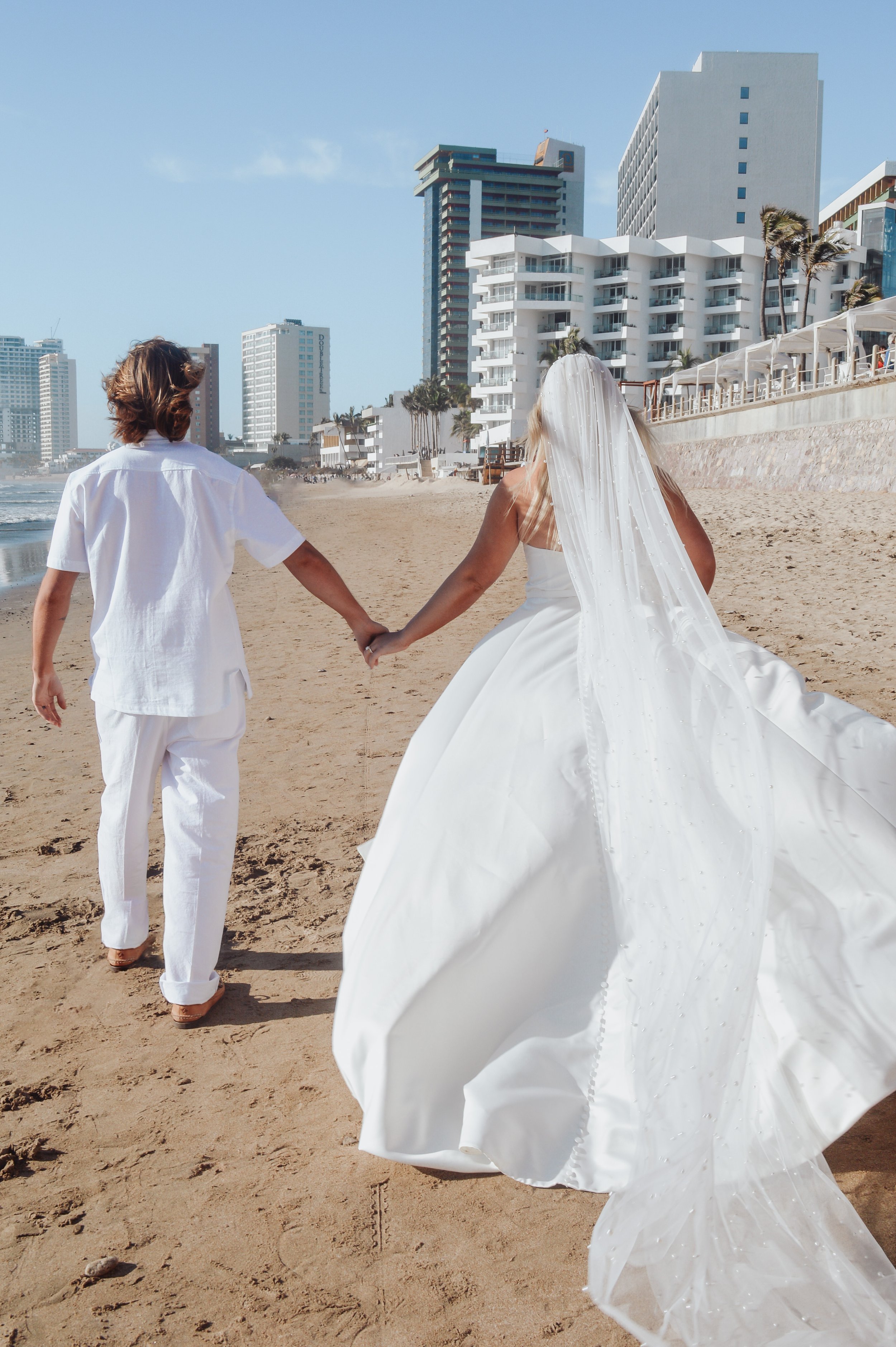 A couple holding hands walking on the beach, the woman is dressed in a wedding gown and veil, and the man is dressed in white linen clothing, with tall buildings and palm trees in the background.
