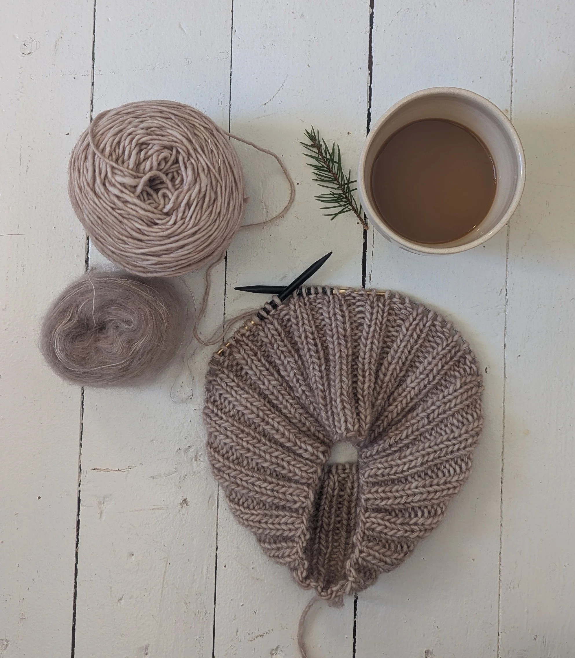 Pöydällä on kaksi kerää lankaa, keskeneräinen neule, virkkuukoukkua, kuppi kahvia ja pieni oksa havupuun neulasta.