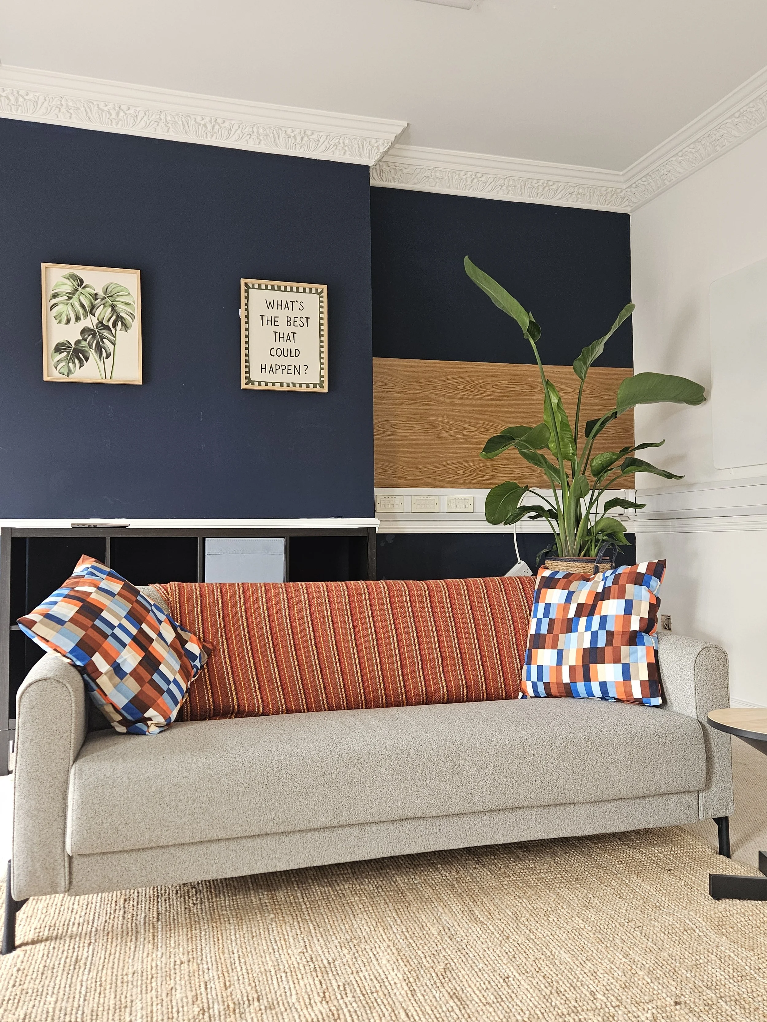 A beige sofa with a plaid-patterned multicolored pillow on each armrest, a red striped throw blanket across the backrest, in a living room with a blue accent wall, framed botanical and motivational quotes, and a large potted plant nearby.