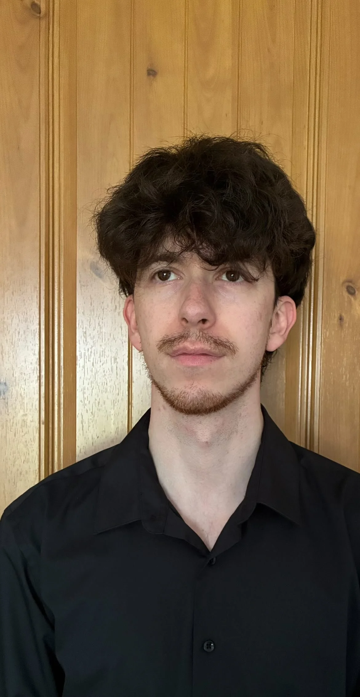A young man with curly dark hair, light skin, and a mustache, wearing a black shirt, standing in front of a wood-paneled wall.