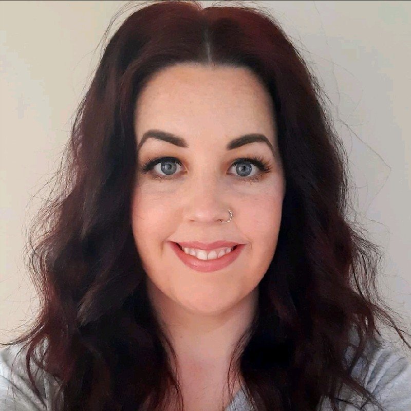 A woman with wavy dark hair and blue eyes, smiling, with a nose ring and a light gray shirt, against a plain background.